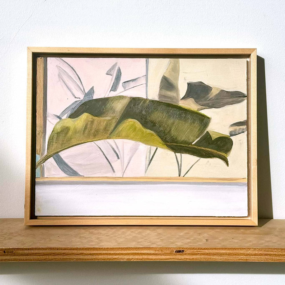 Framed oil painting artwork with a large green leaf on a wooden shelf against a white wall