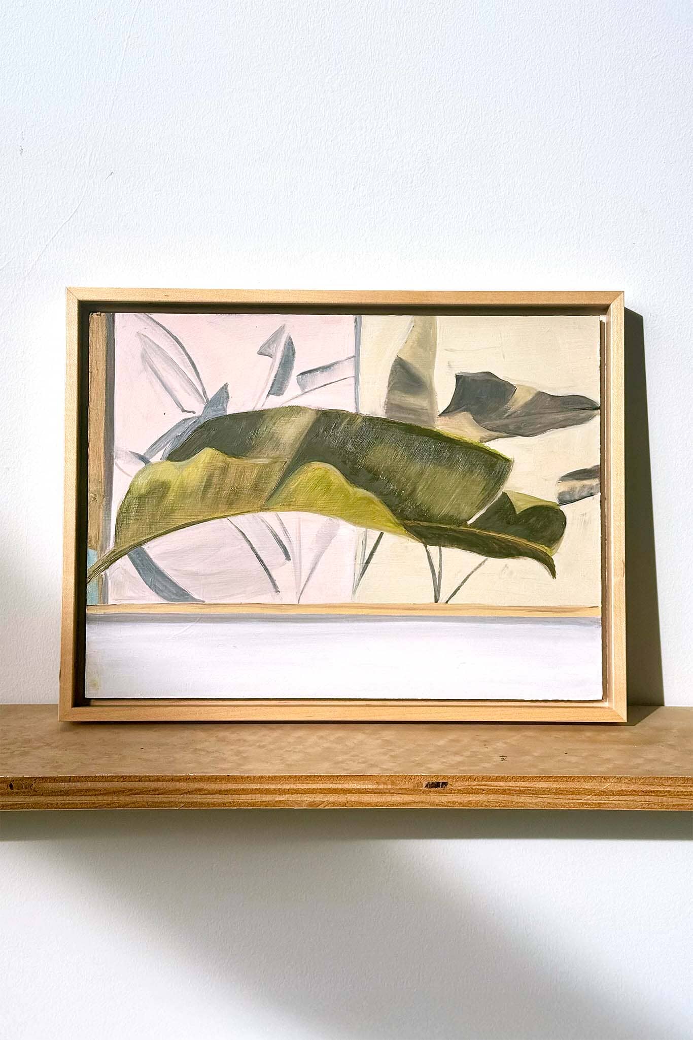 Framed oil painting artwork with a large green leaf on a wooden shelf against a white wall