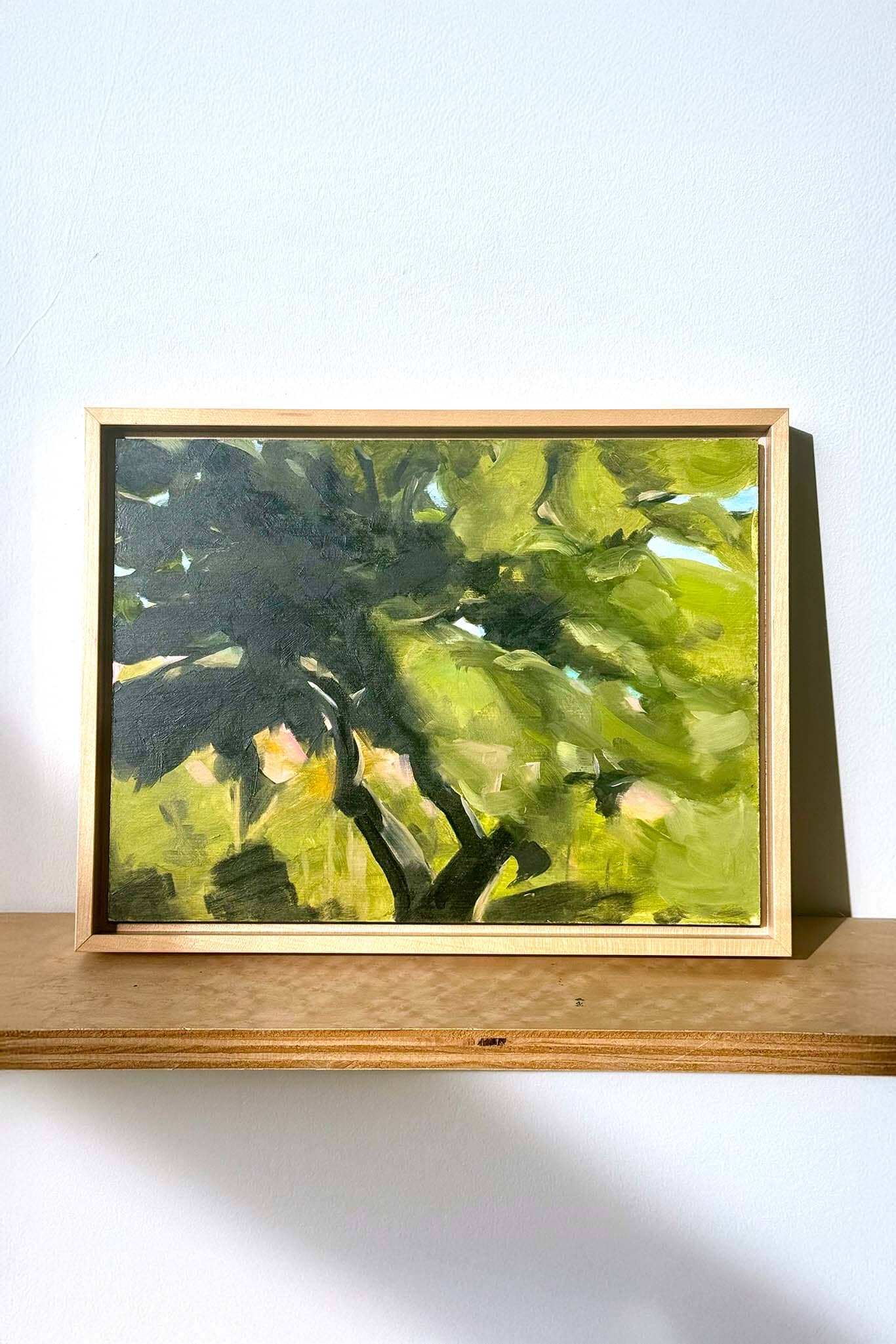 Framed painting of a green leafy scene on a wooden shelf with a white background