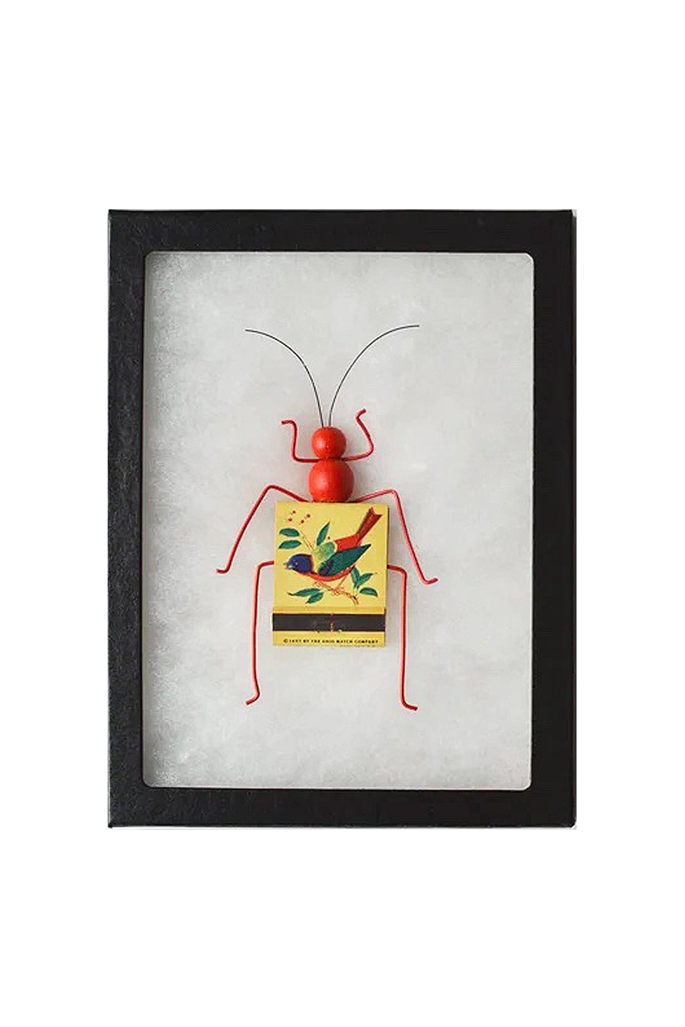 Framed artwork of a red insect-like figure with a yellow matchbook featuring a bird design on a white background.