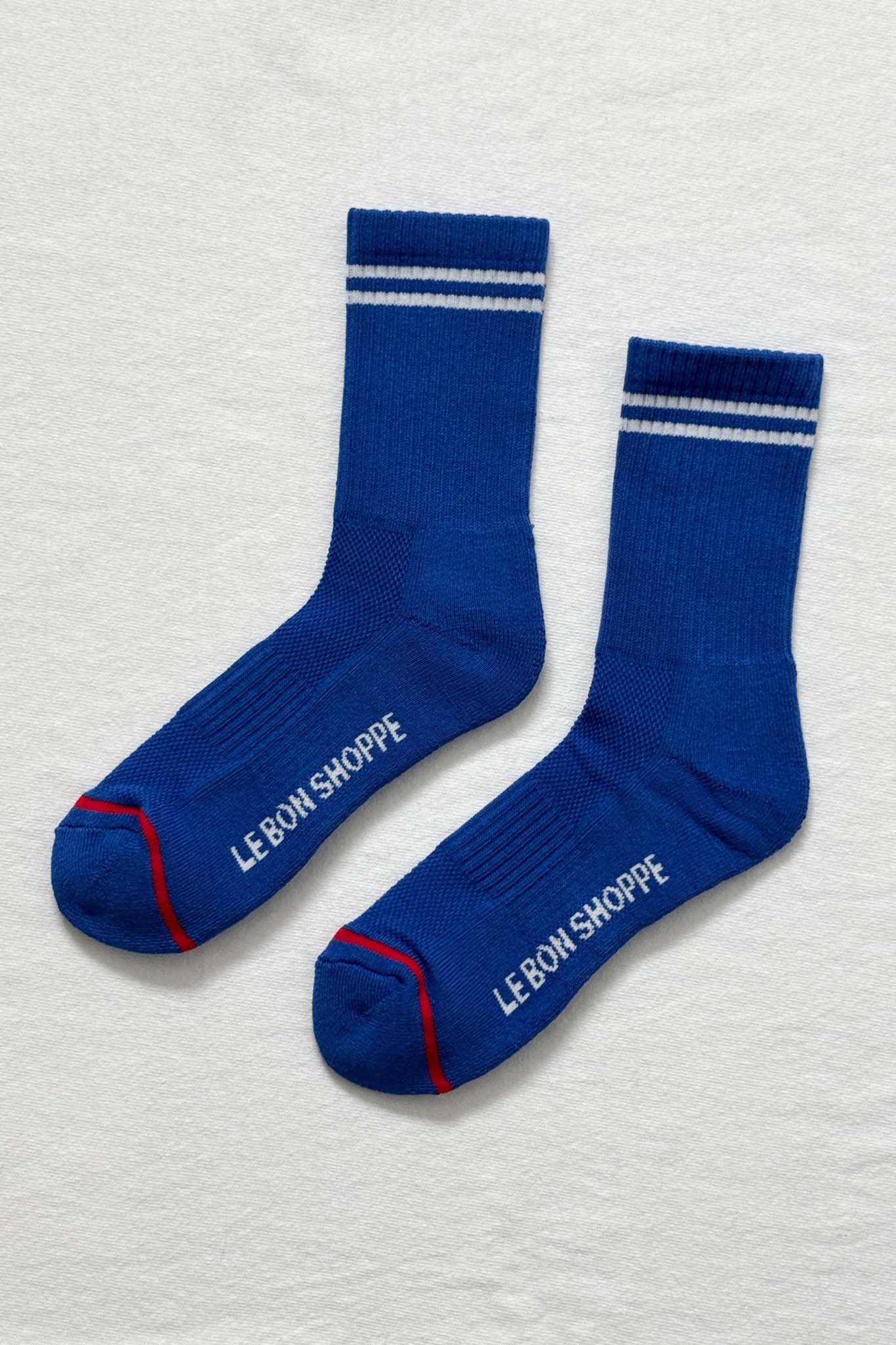 Pair of blue socks with white stripes and 'Le Bon Shoppe' branding on a light gray background