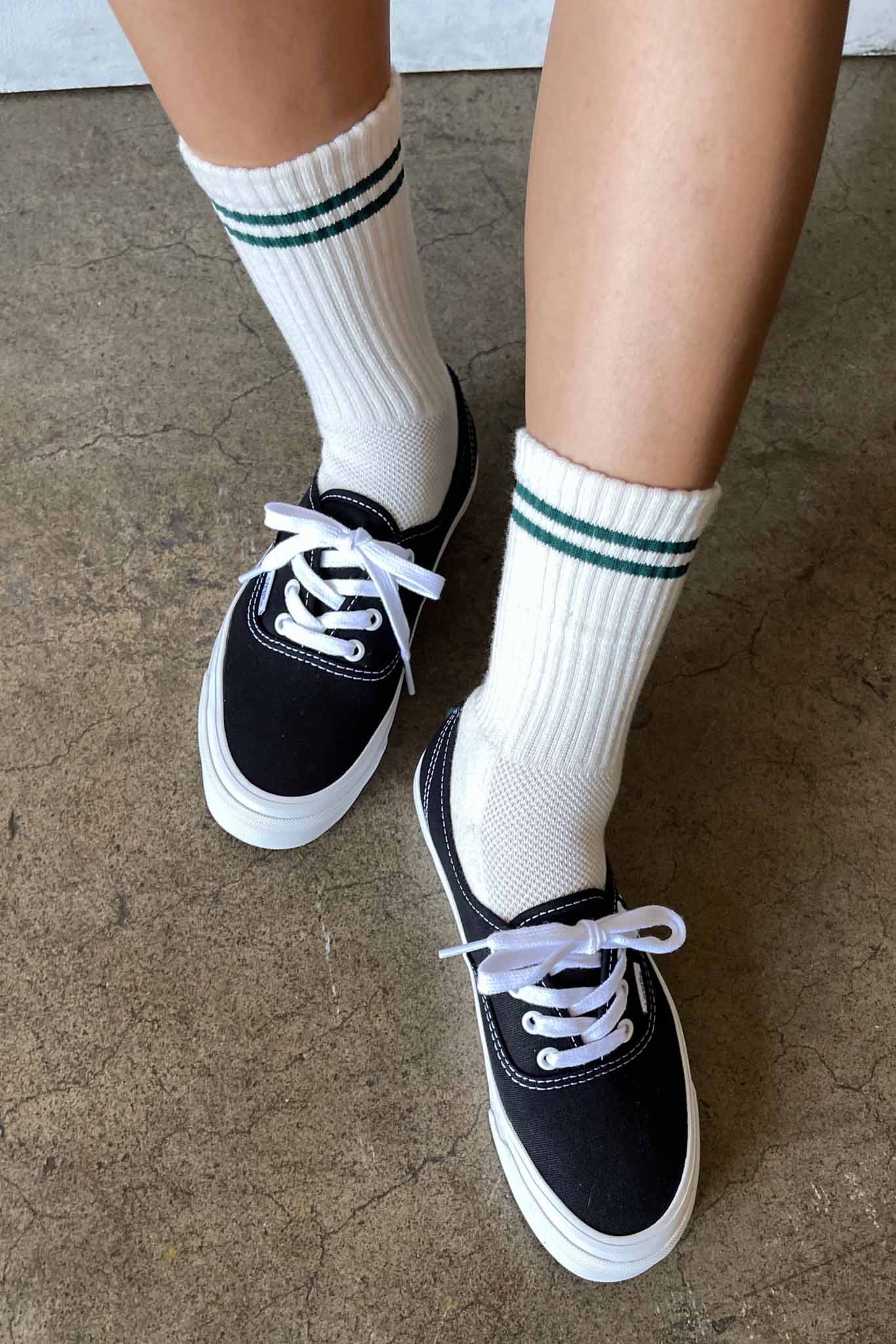 Black sneakers with white laces worn with white socks featuring green stripes on a concrete floor.