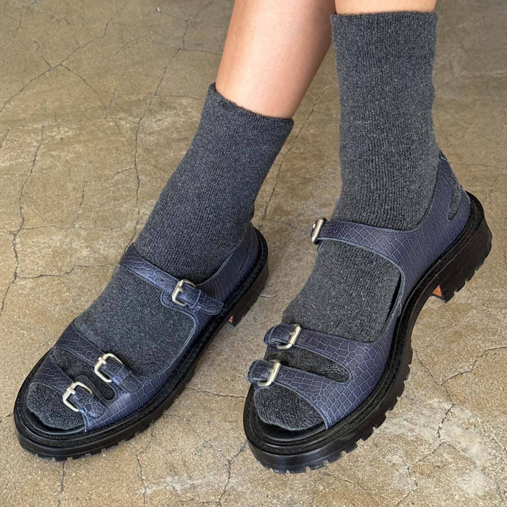 Dark blue sandals with buckles worn with gray socks on a stone floor.