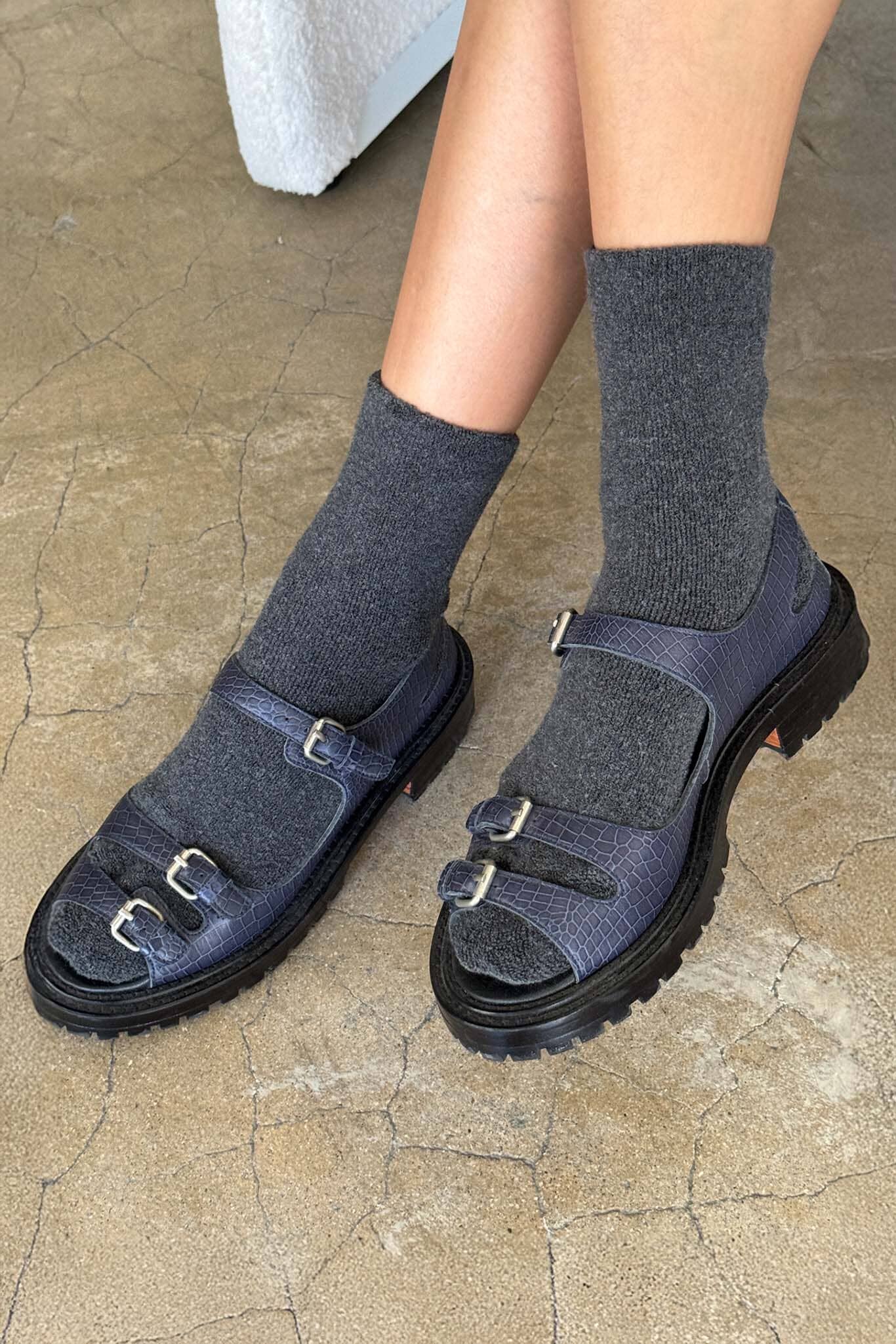 Dark blue sandals with buckles worn with gray socks on a stone floor.