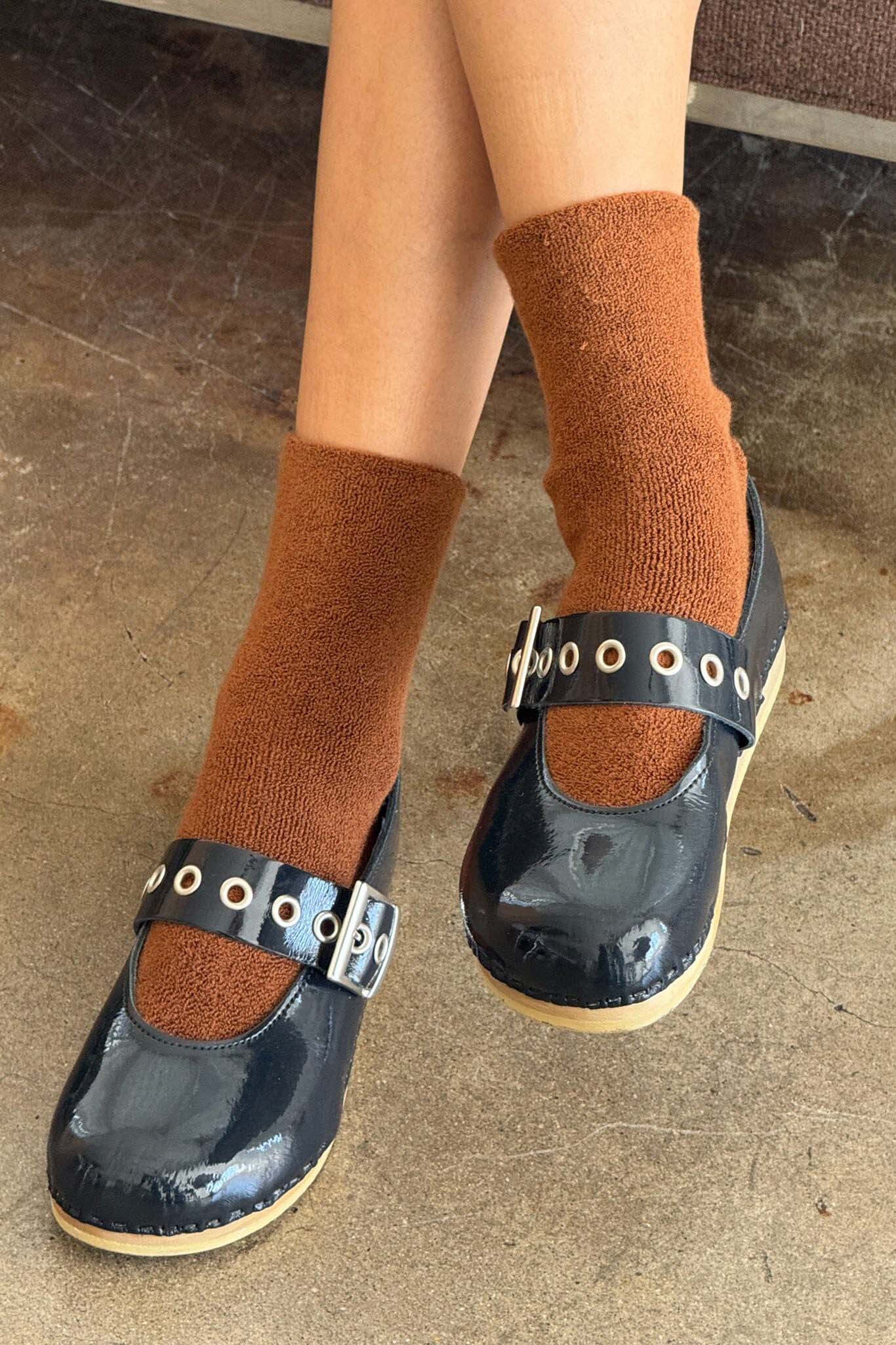 Black shoes with buckle details worn with brown socks on a concrete floor.