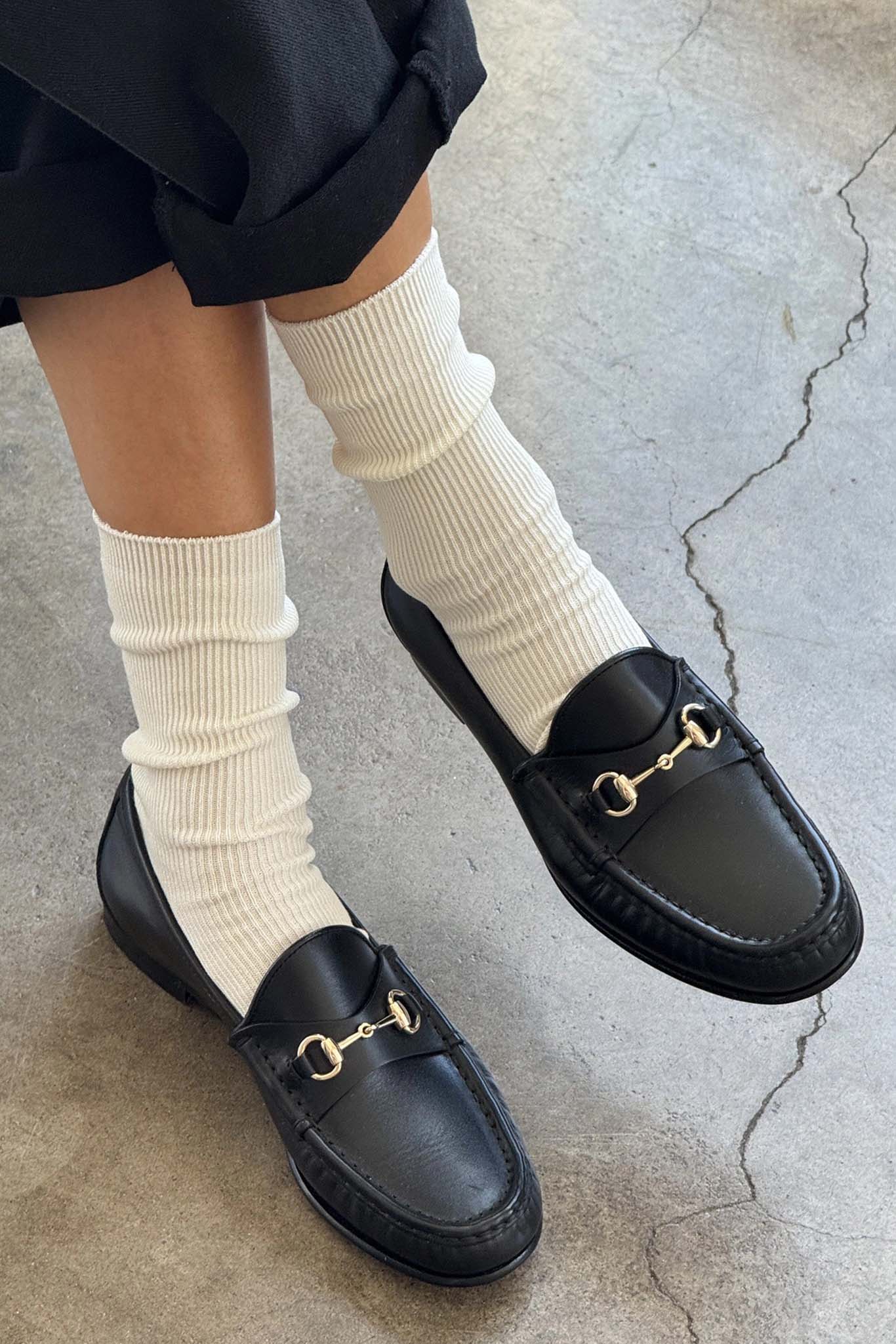 Black loafers with gold buckles worn with white socks on a concrete surface