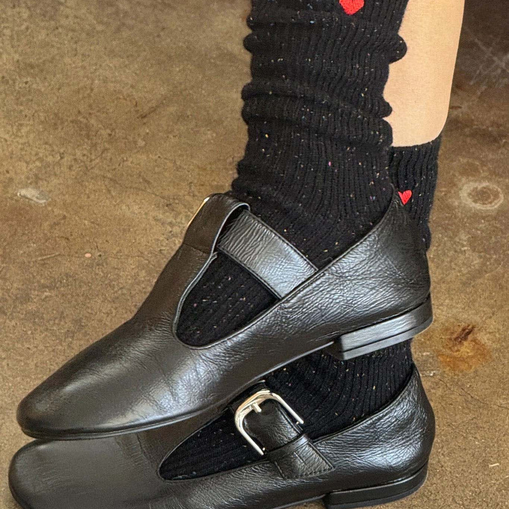 Black leather shoes with a strap and buckle worn with black socks featuring red hearts on a concrete floor.