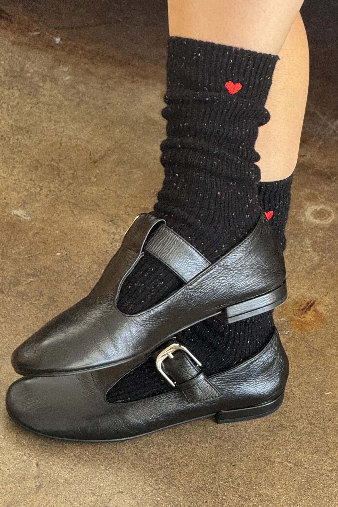 Black leather shoes with a strap and buckle worn with black socks featuring red hearts on a concrete floor.