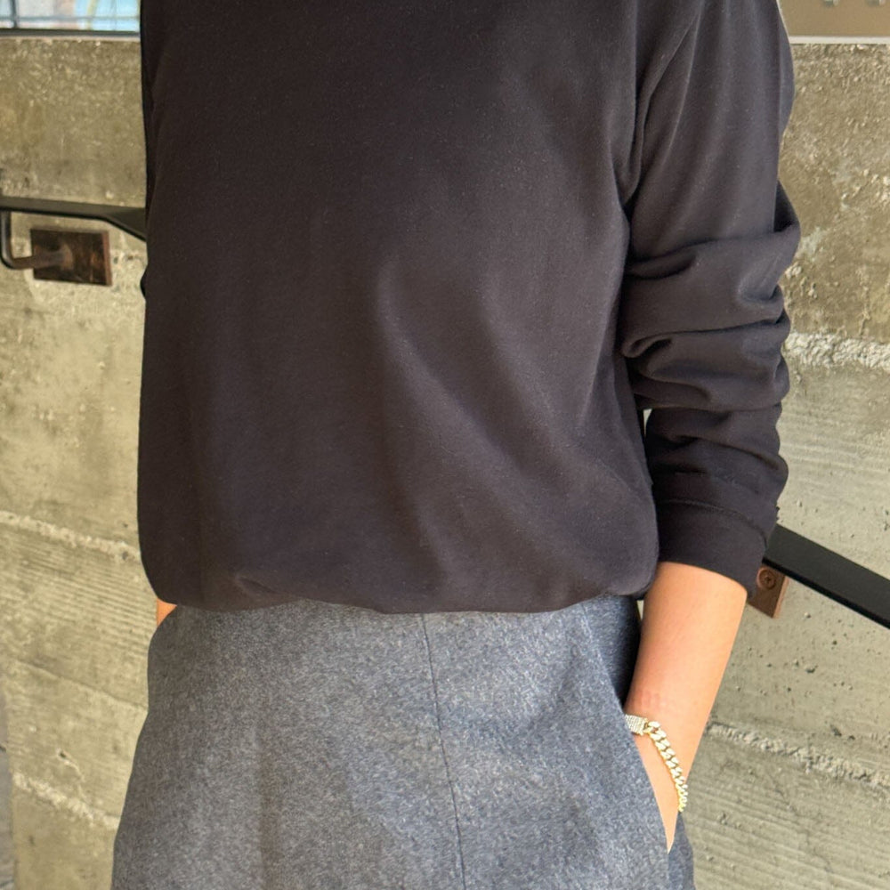 Person wearing a black long-sleeve top and gray skirt standing against a concrete wall.