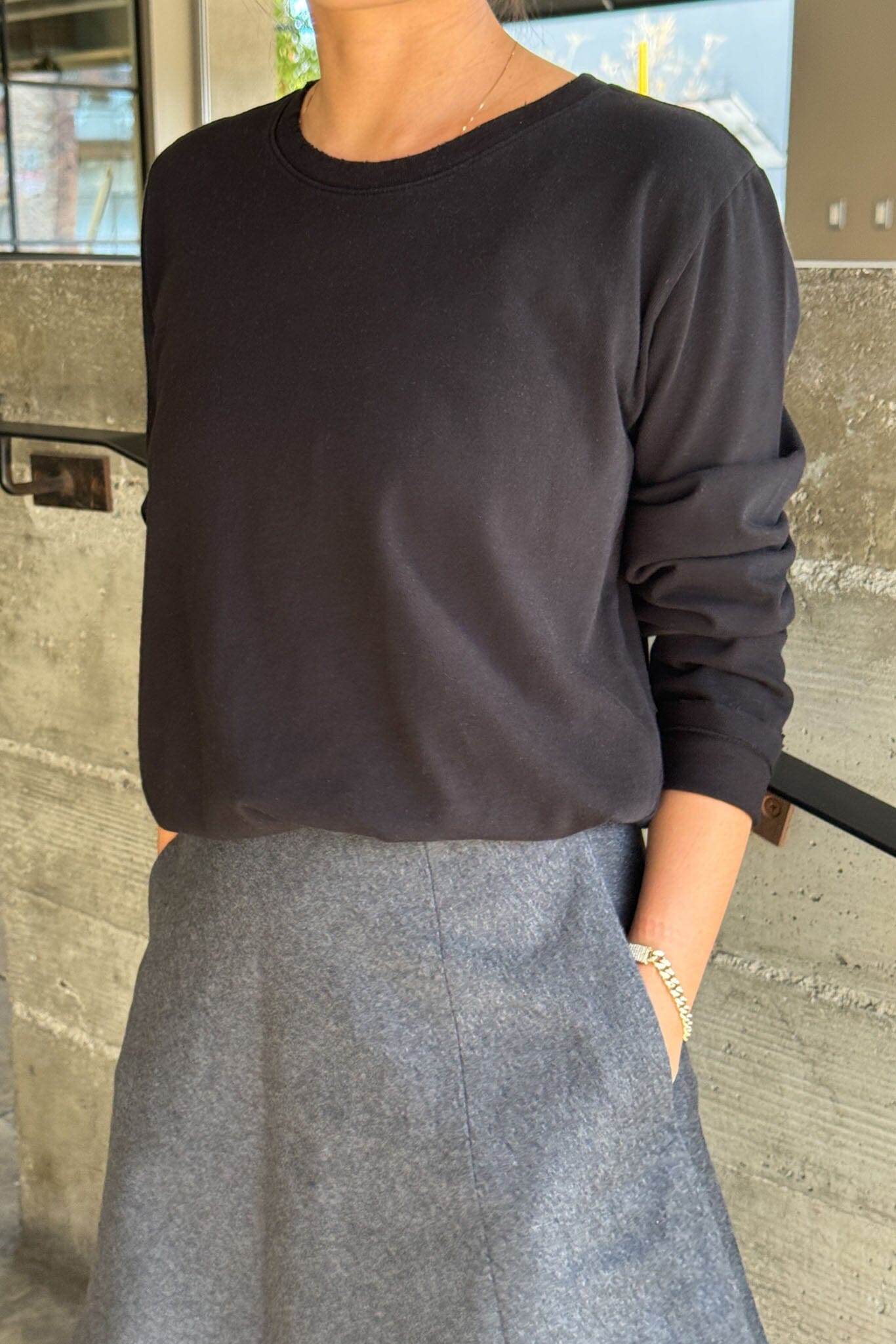 Person wearing a black long-sleeve top and gray skirt standing against a concrete wall.