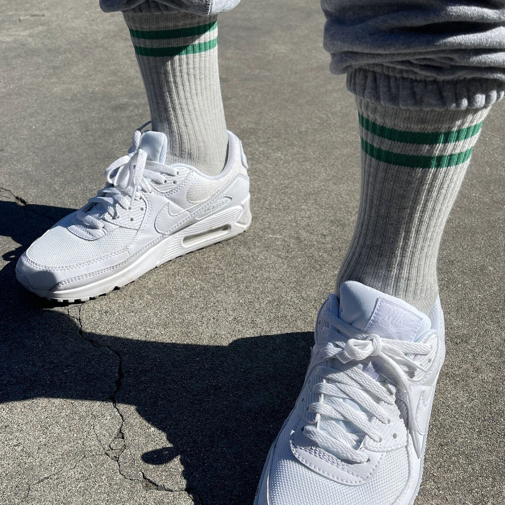 Sneakers worn with gray sweatpants and white socks with green stripes on a concrete surface.