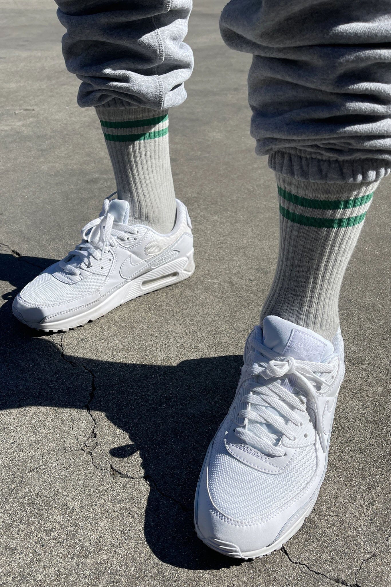 Sneakers worn with gray sweatpants and white socks with green stripes on a concrete surface.