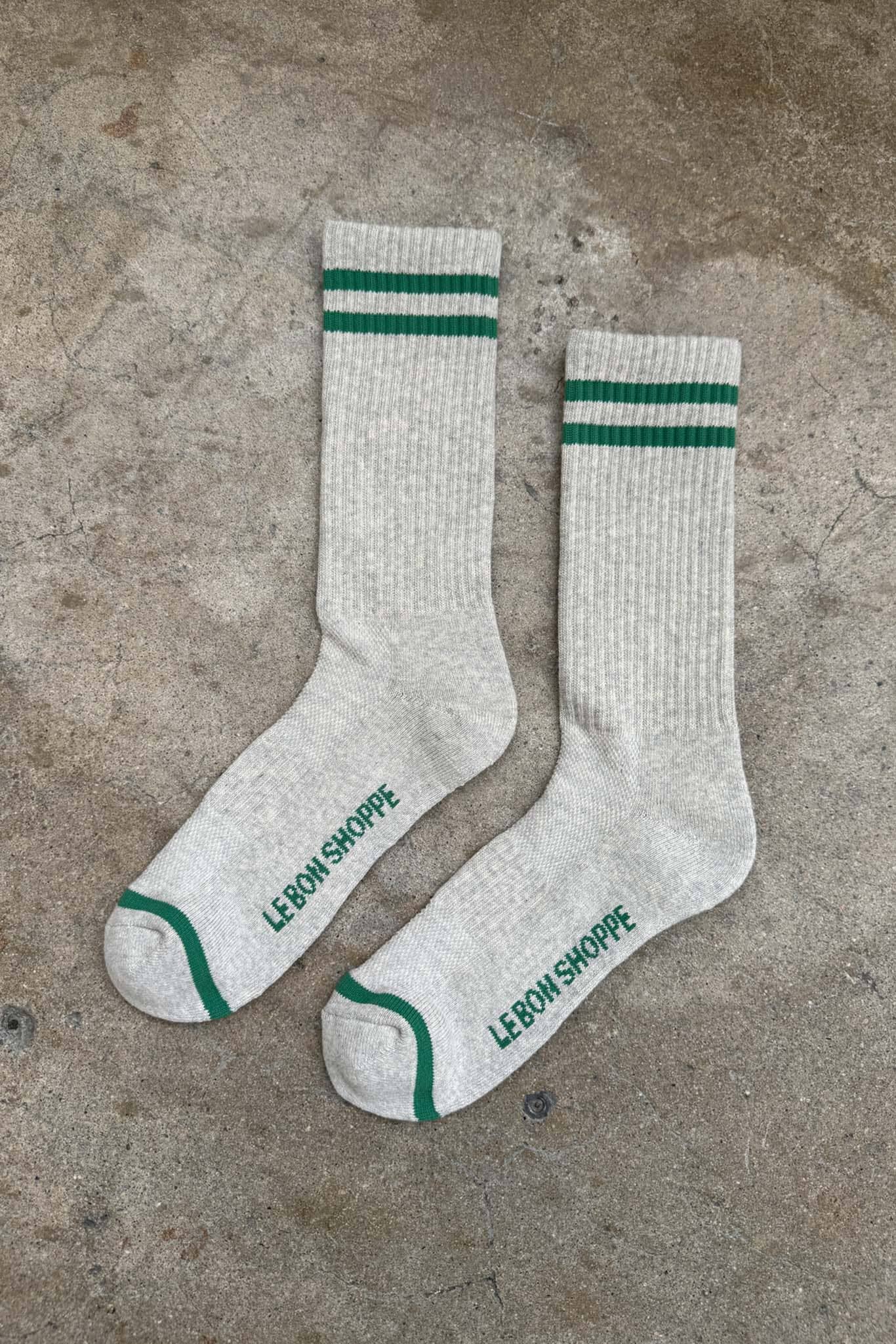 Two white socks with green stripes and 'Le Bon Shoppe' branding on a concrete surface.