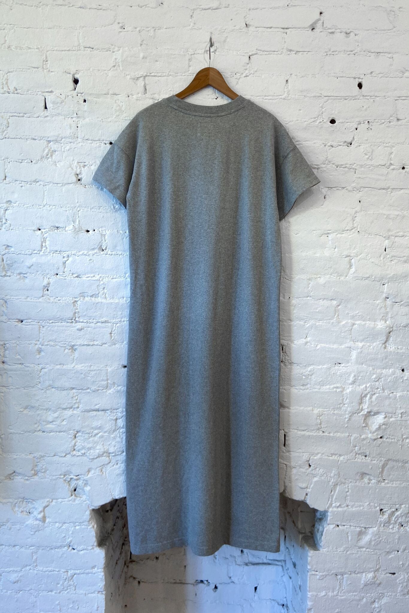 A light gray short-sleeve knit dress or tunic top hanging on a wooden hanger against a white painted brick wall.