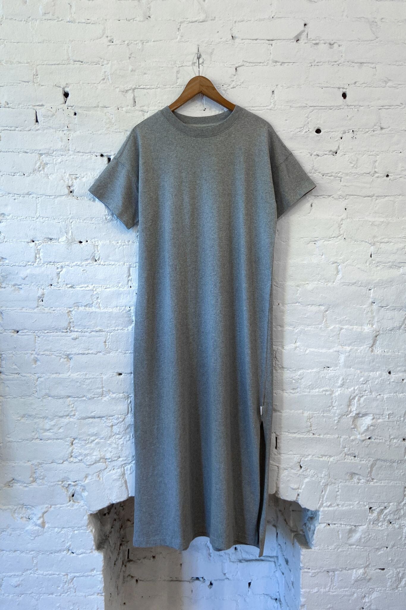 A short-sleeve crewneck knit dress in heather gray, hanging on a wooden hanger against a white brick wall.