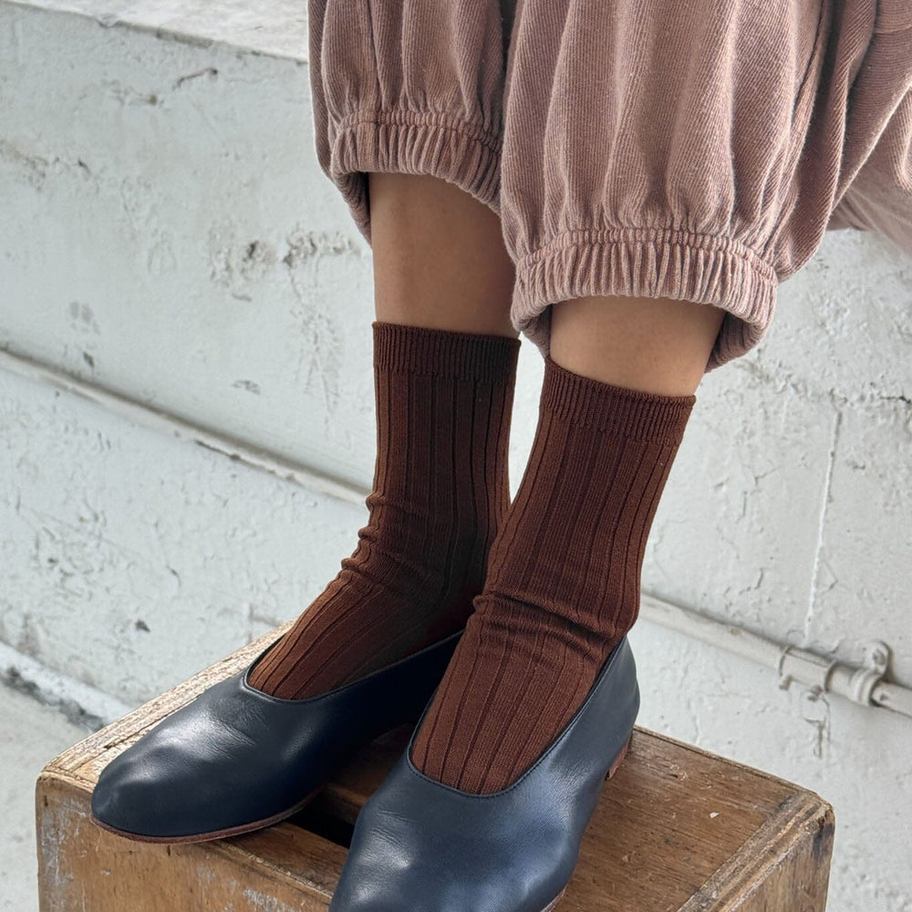 Person wearing brown socks and navy flats on a wooden box with a light-colored wall background