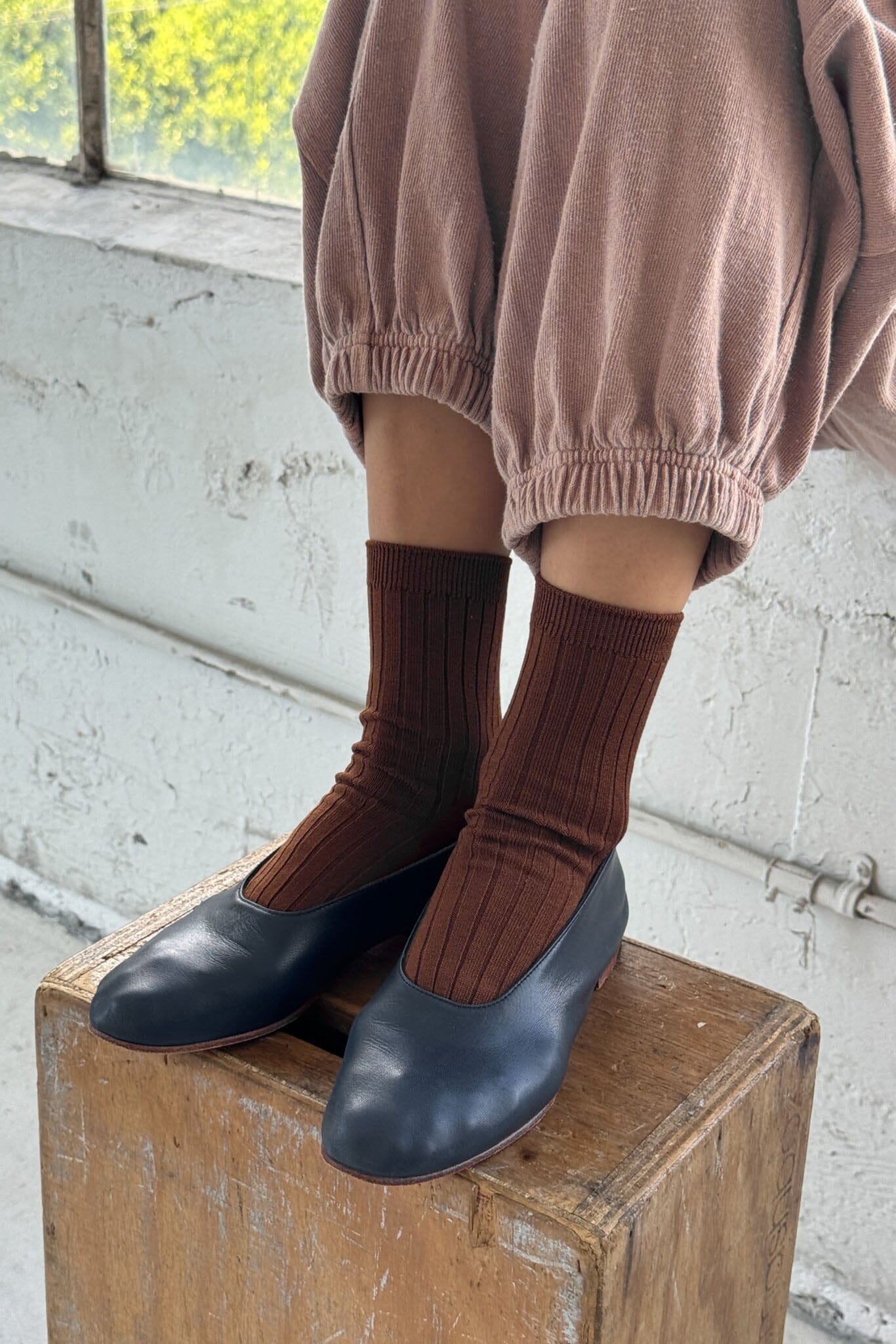 Person wearing brown socks and navy flats on a wooden box with a light-colored wall background