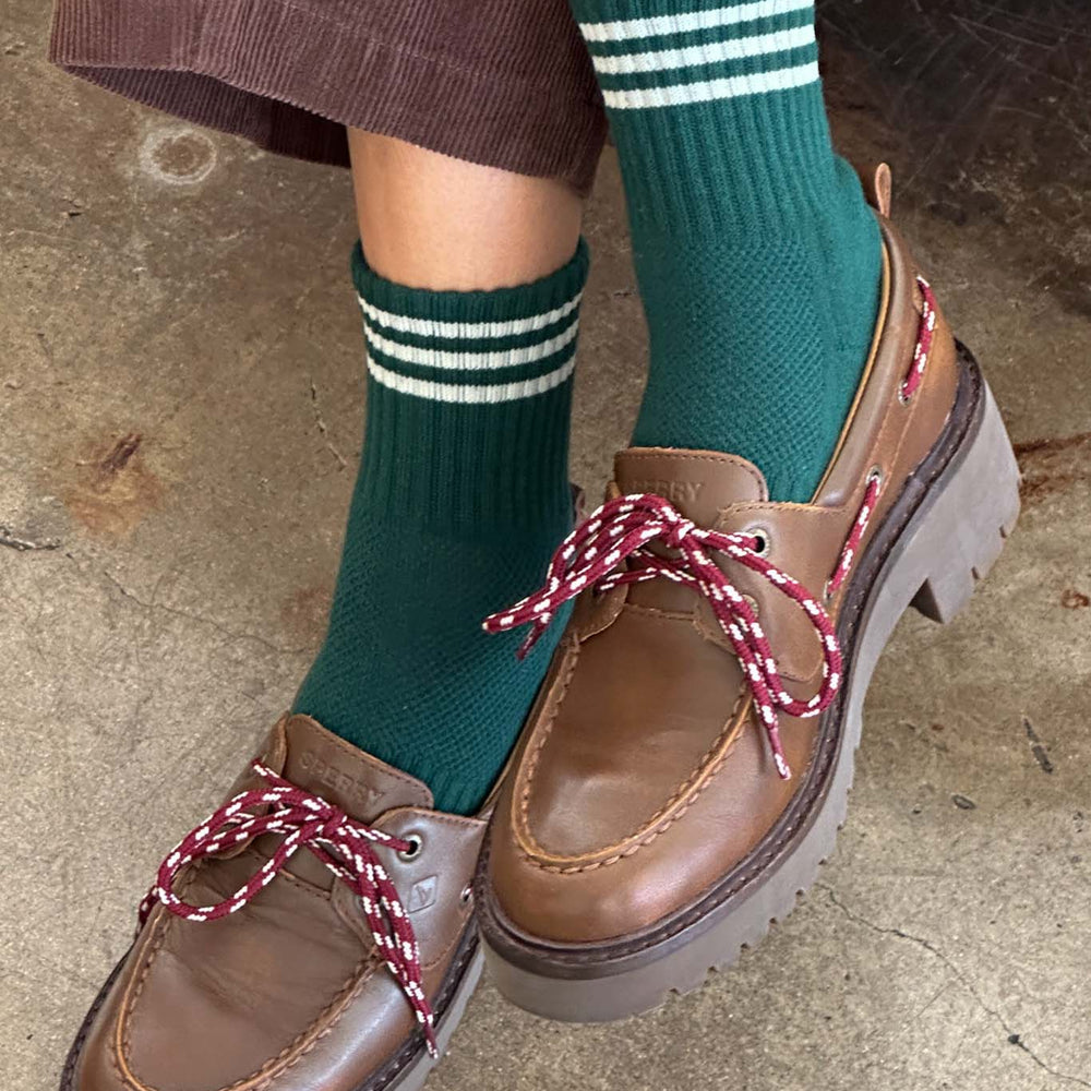Brown loafers with red laces worn with green striped crew socks on a concrete floor.