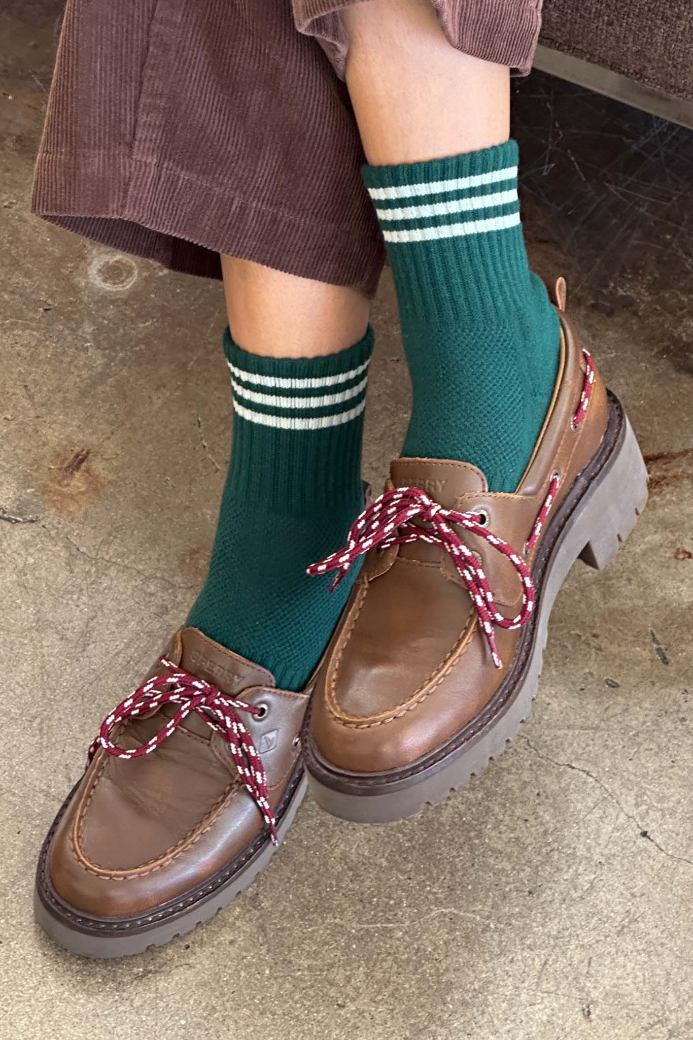 Brown loafers with red laces worn with green striped crew socks on a concrete floor.