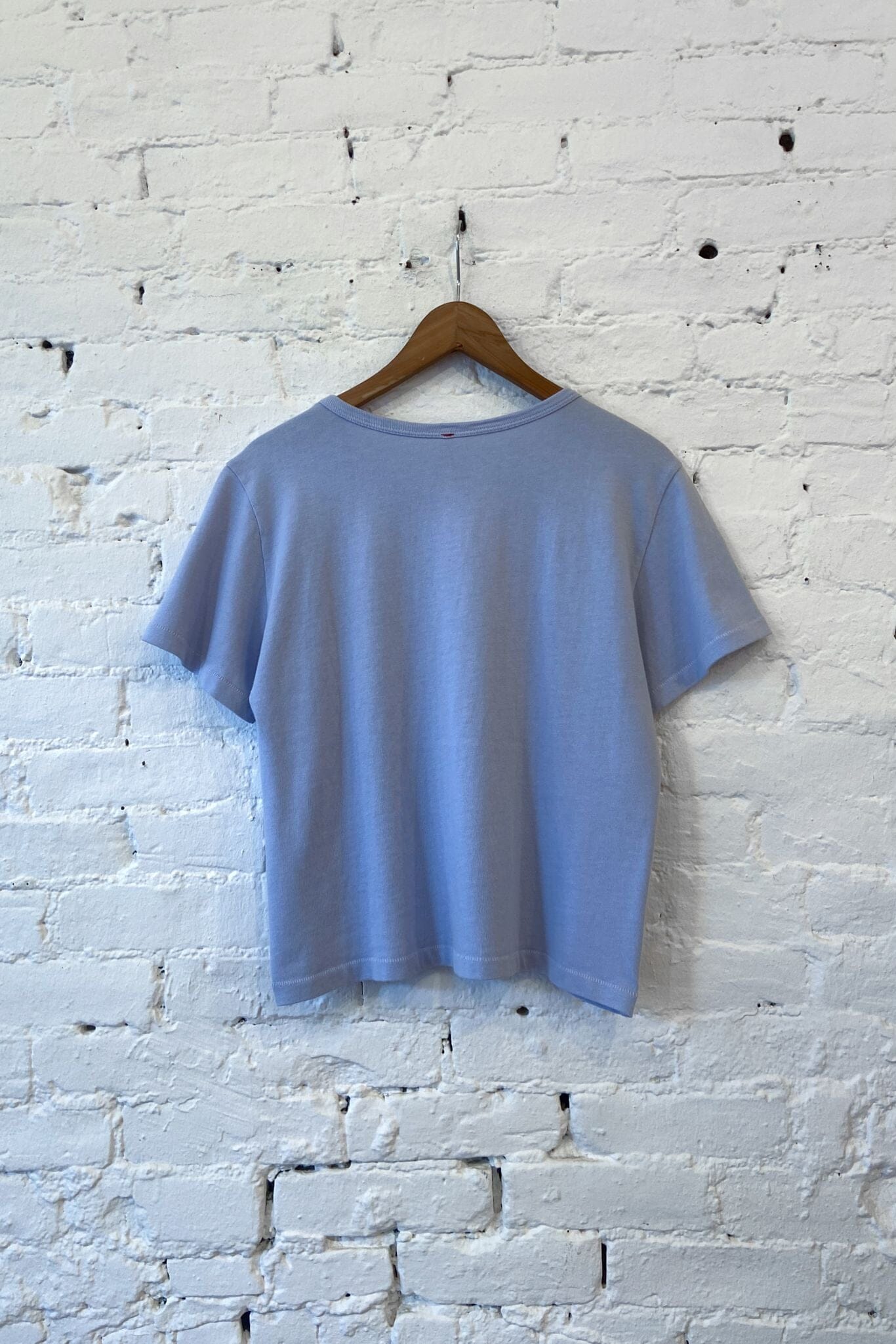 A light periwinkle blue short-sleeve t-shirt hanging on a wooden hanger against a white-painted brick wall.