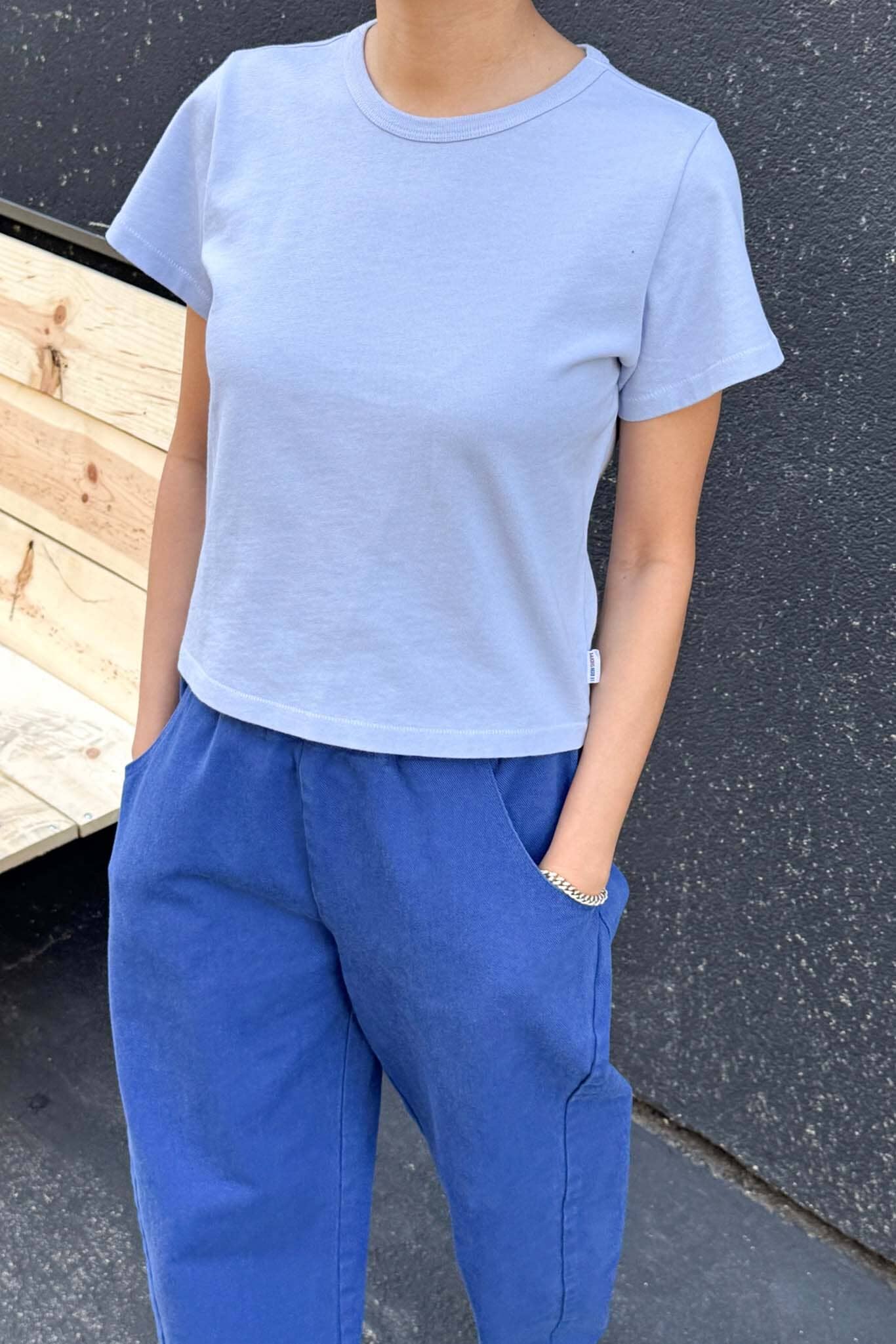 Person wearing a light blue t-shirt and blue pants against a dark wall.