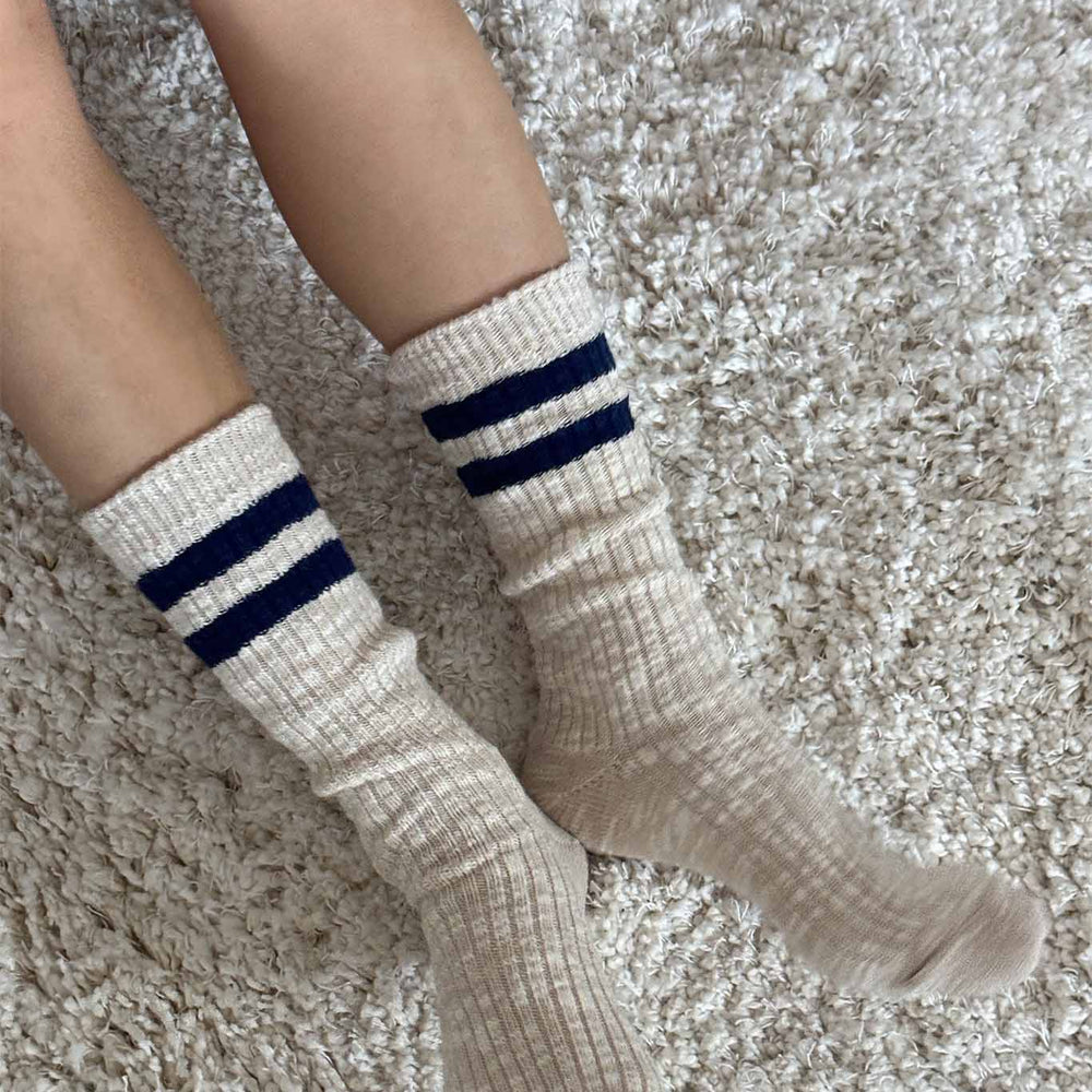 Beige ribbed socks with two blue stripes worn by a person on a textured carpet.