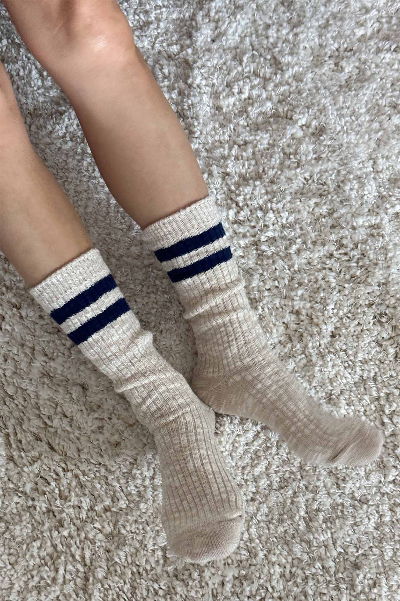 Beige ribbed socks with two blue stripes worn by a person on a textured carpet.