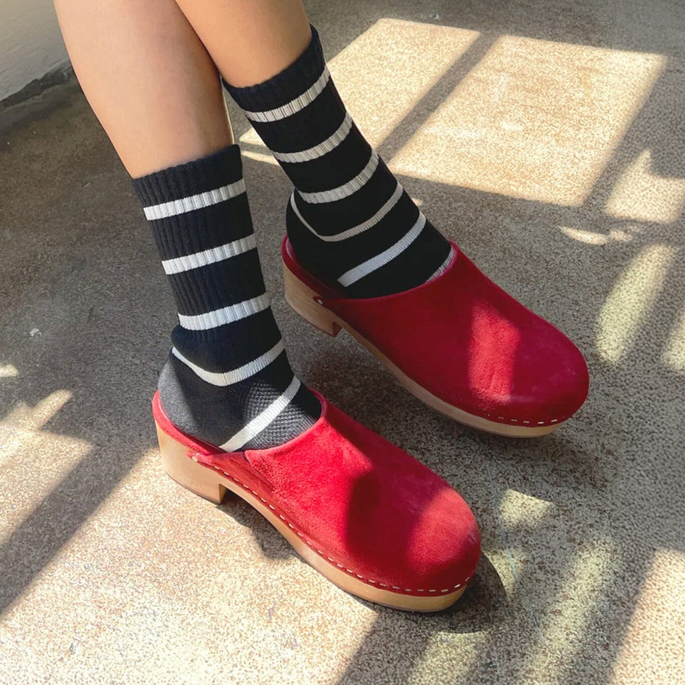 A person wearing black and white striped socks and red clogs standing on concrete ground with shadows cast over the scene.