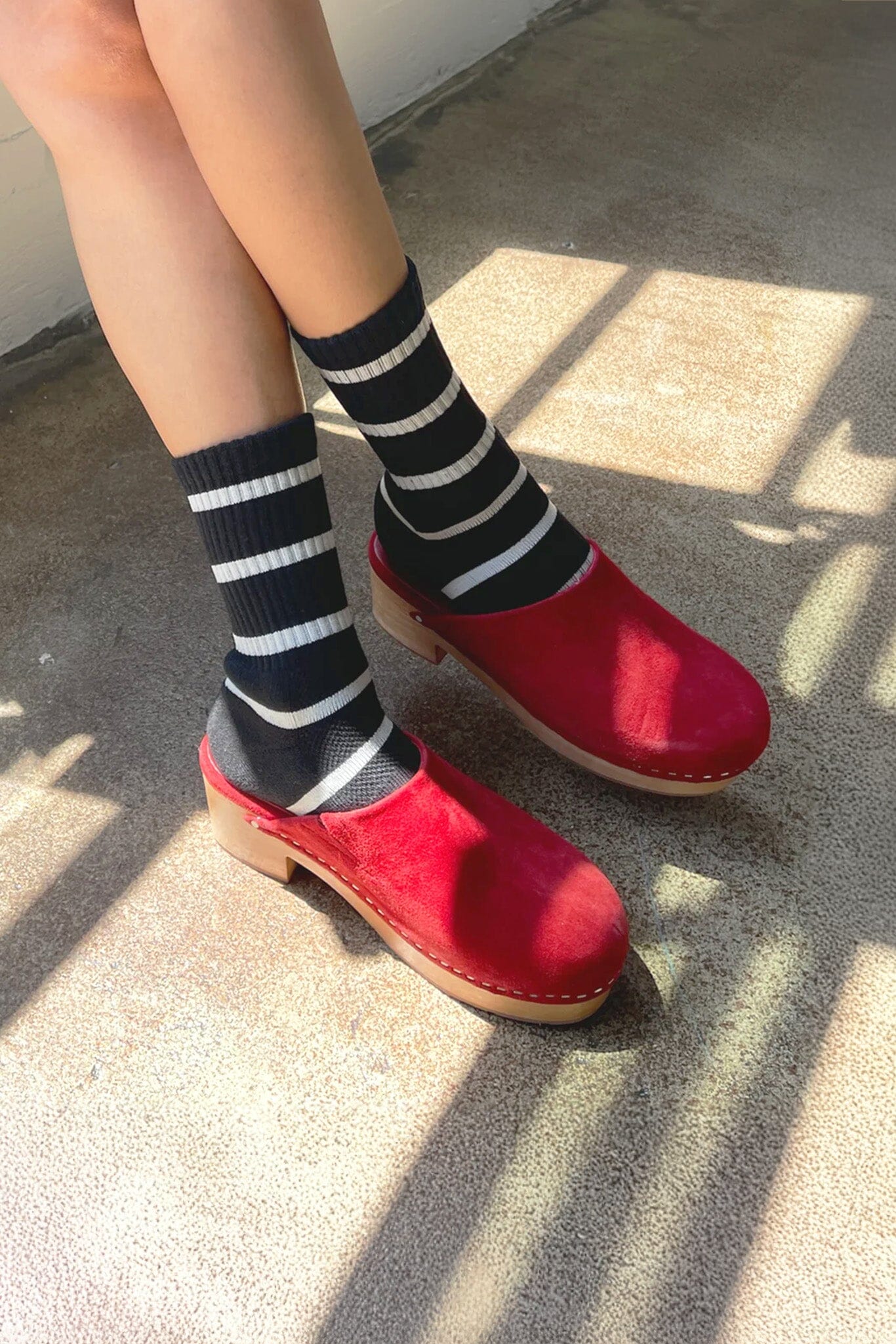 A person wearing black and white striped socks and red clogs standing on concrete ground with shadows cast over the scene.