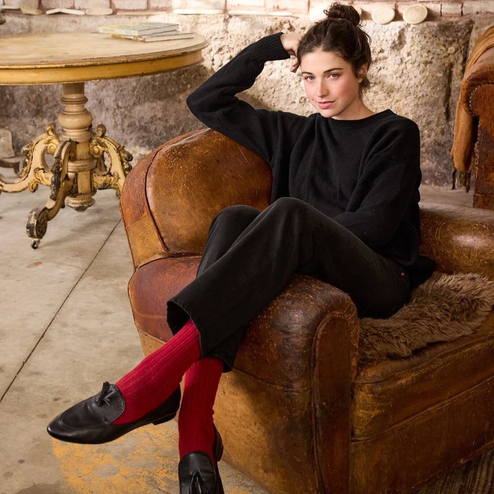 Woman in black outfit with ribbed cashmere socks sitting on a brown leather chair in a rustic setting.