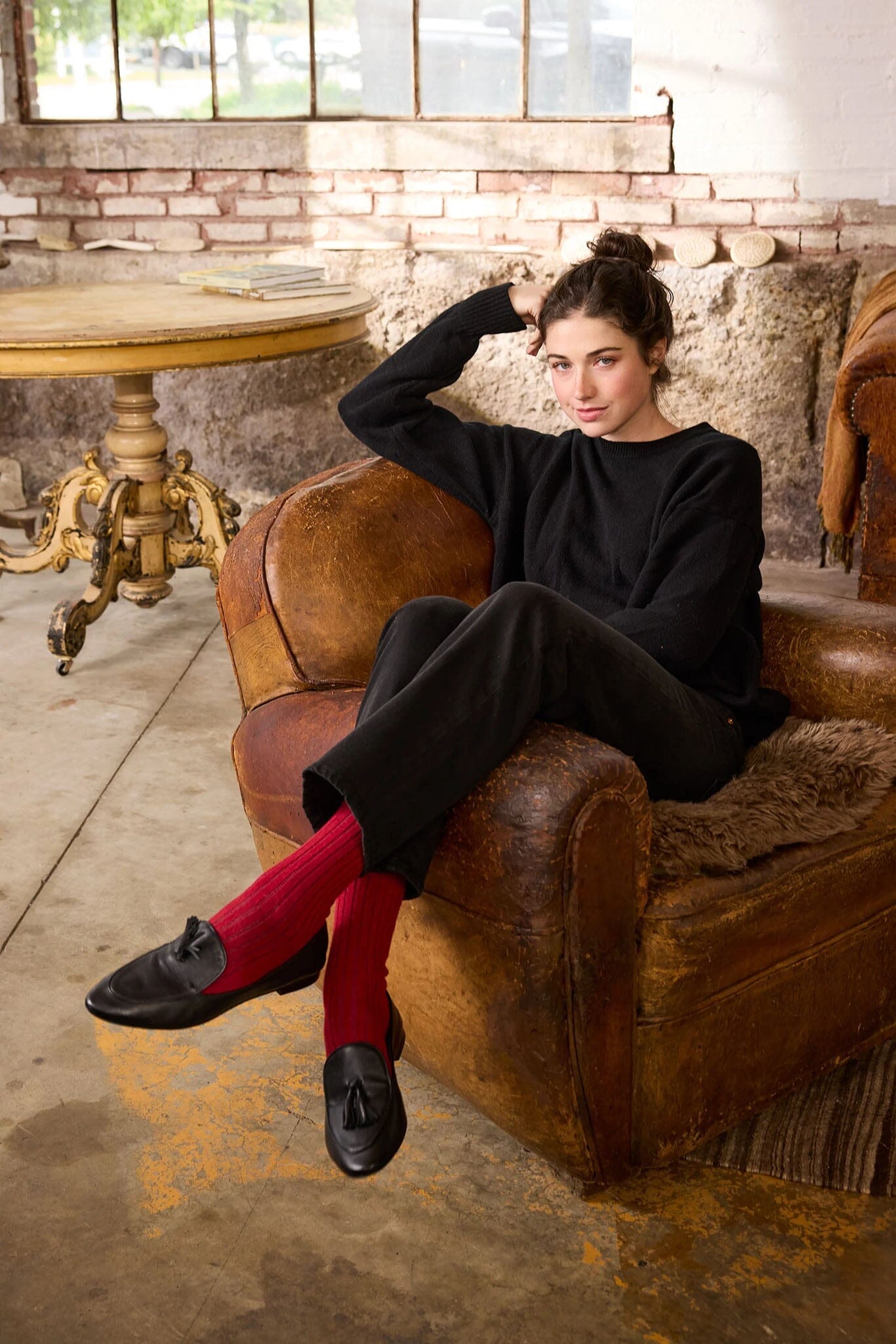 Woman in black outfit with ribbed cashmere socks sitting on a brown leather chair in a rustic setting.
