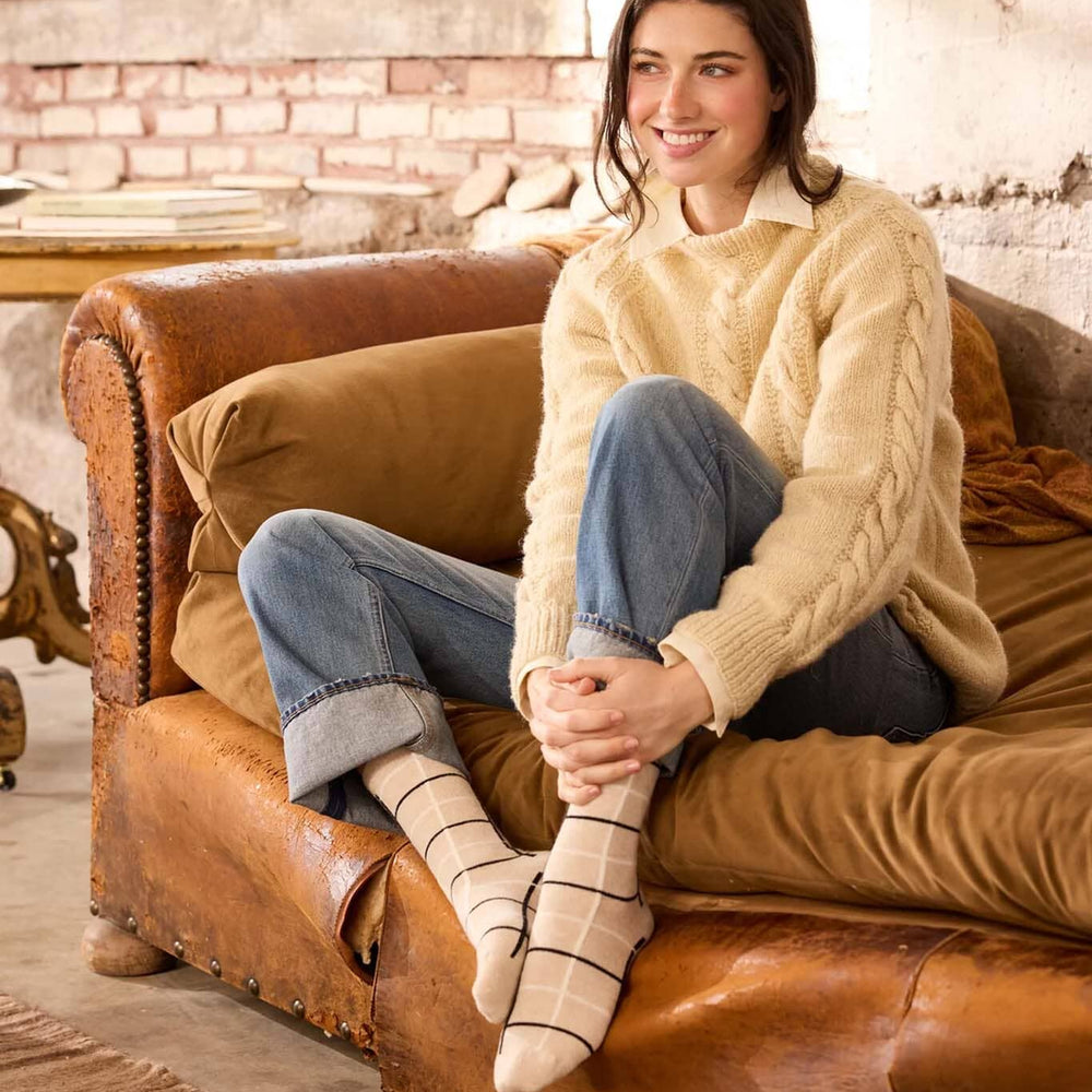 Woman sitting on a brown leather couch wearing a beige sweater and blue jeans.