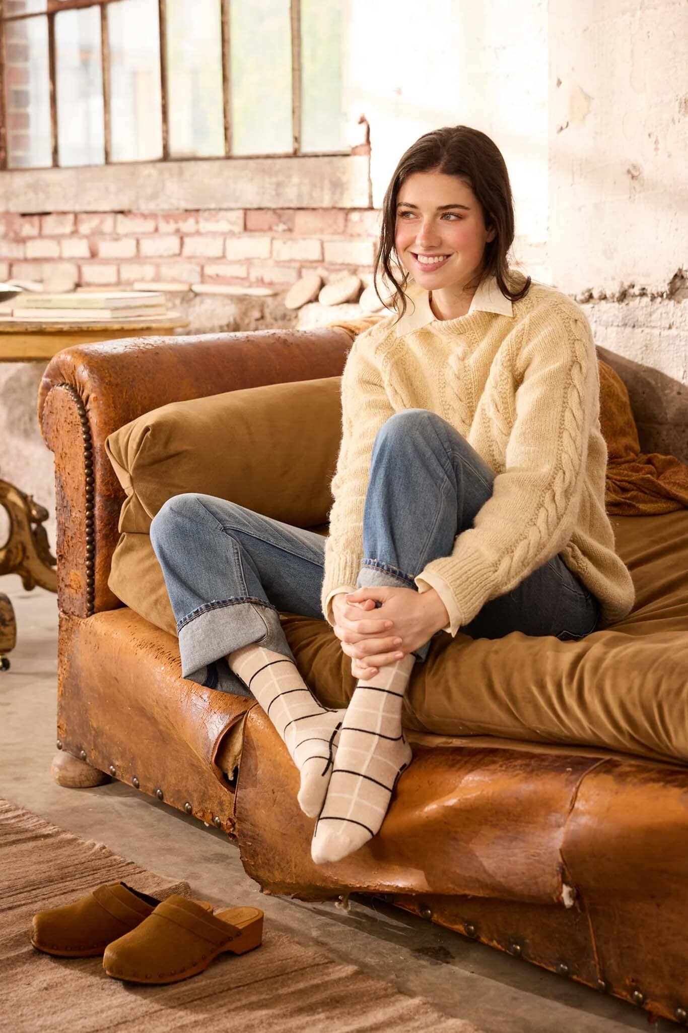 Woman sitting on a brown leather couch wearing a beige sweater and blue jeans.