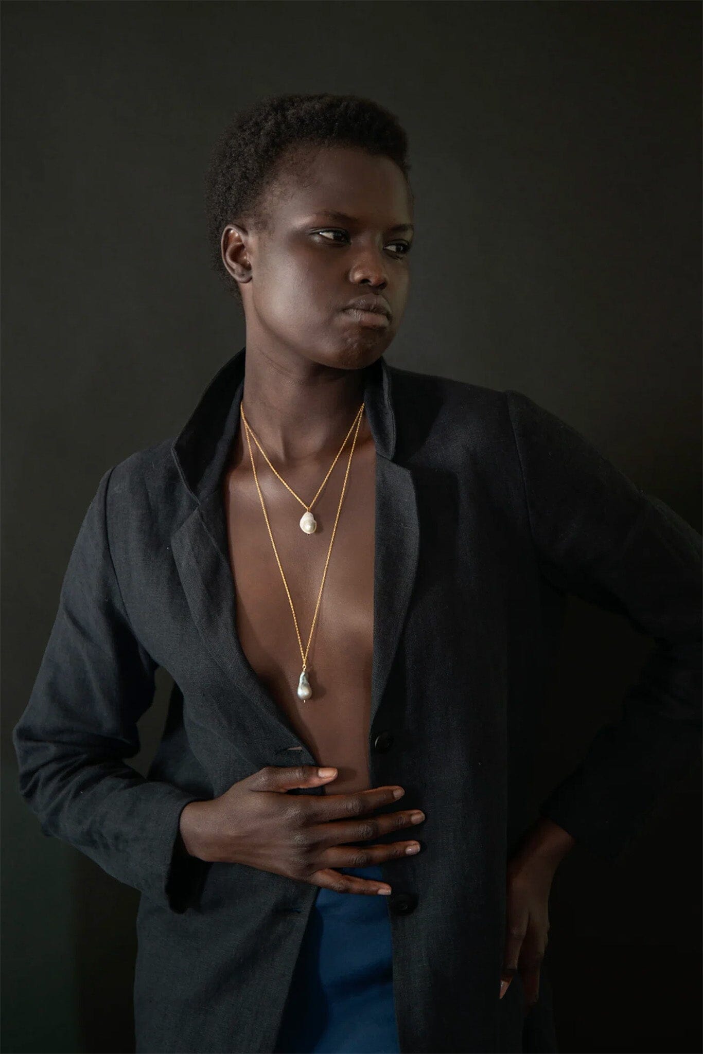 Person wearing a dark jacket over a blue garment with gold necklaces against a dark background