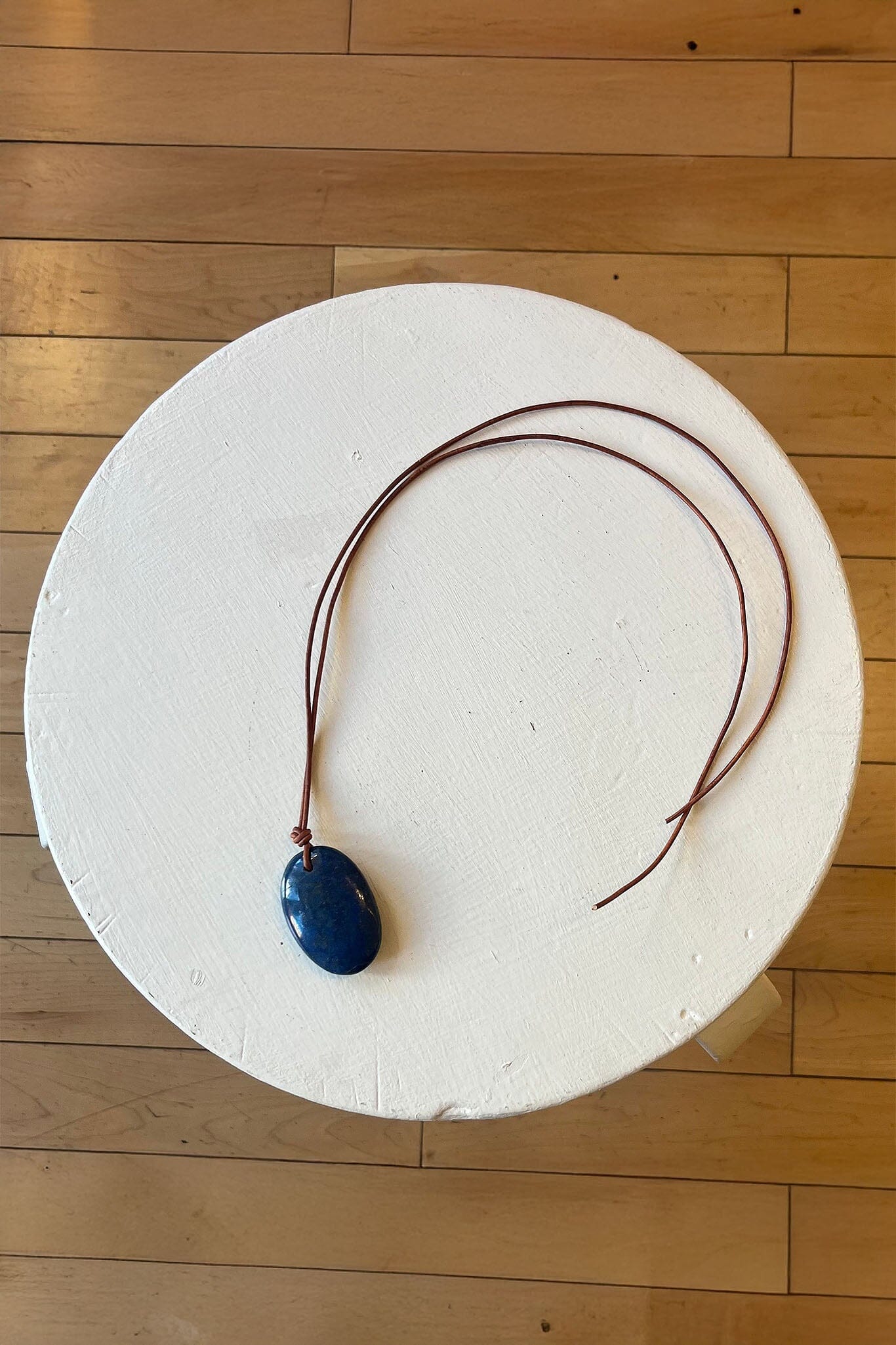 Necklace with an oval blue lapis pendant on a white circular stand against a wooden floor background