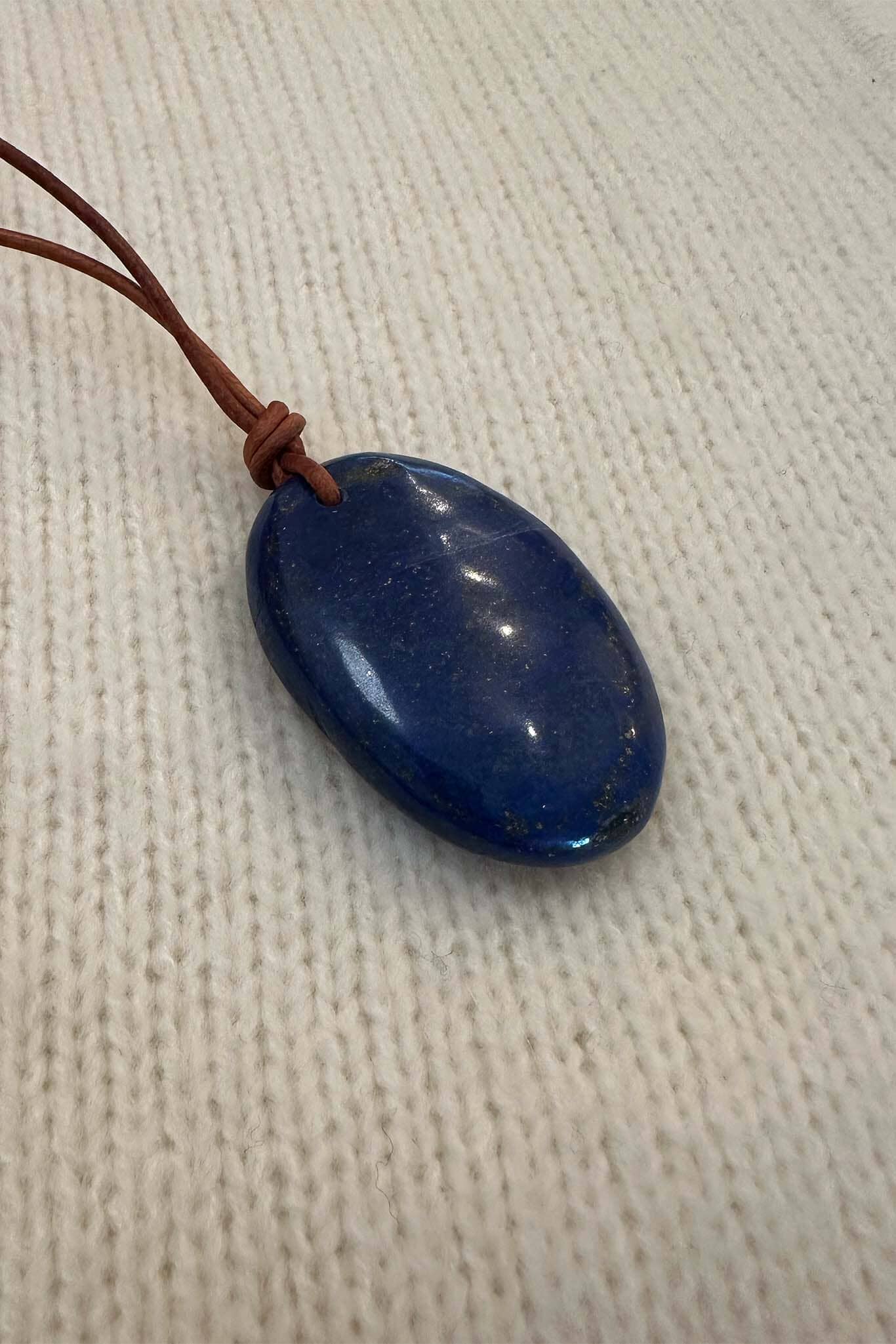 Blue oval lapis pendant on a brown cord against a beige fabric background