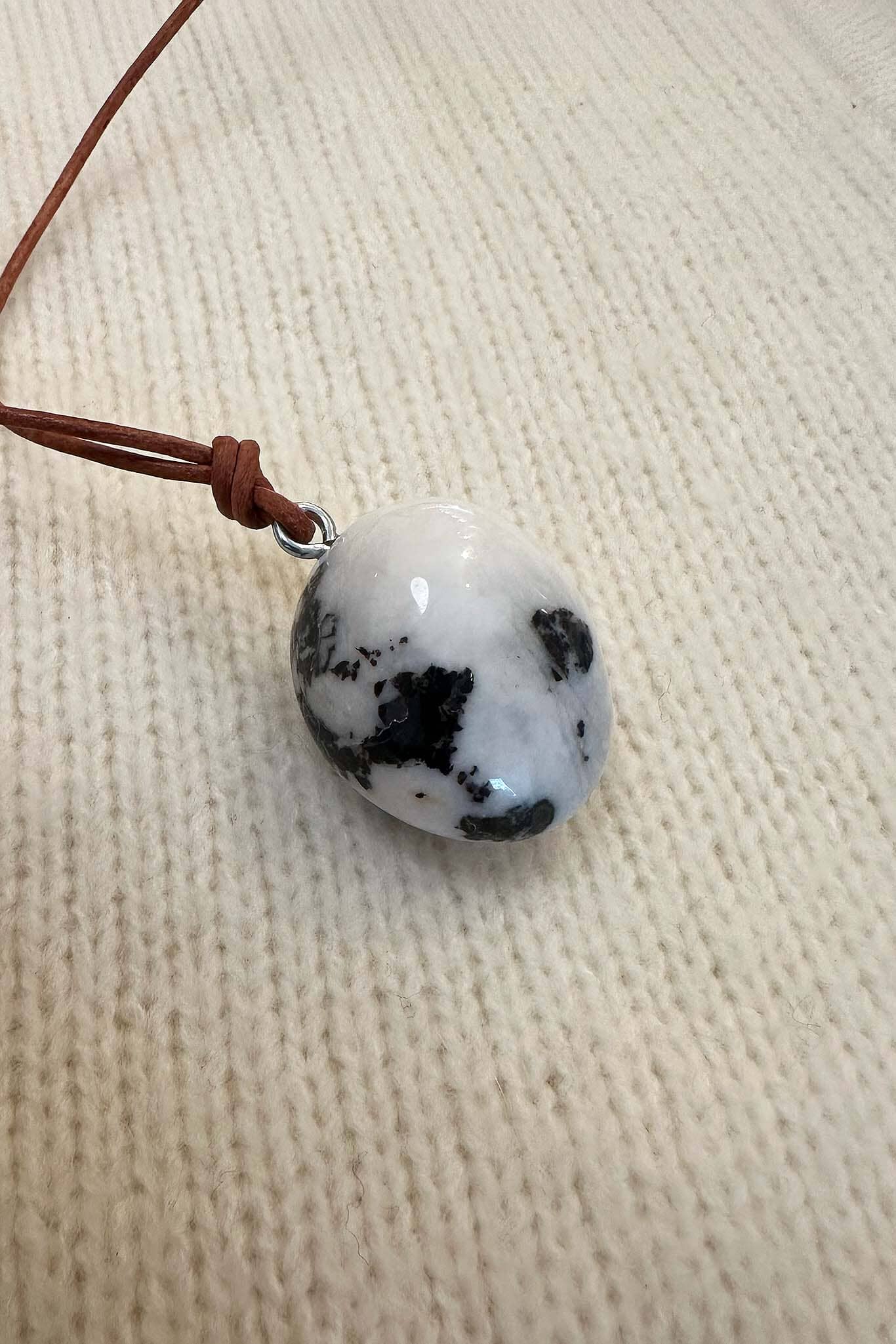 Round white stone pendant with black speckles on a brown cord, lying on a textured beige surface.