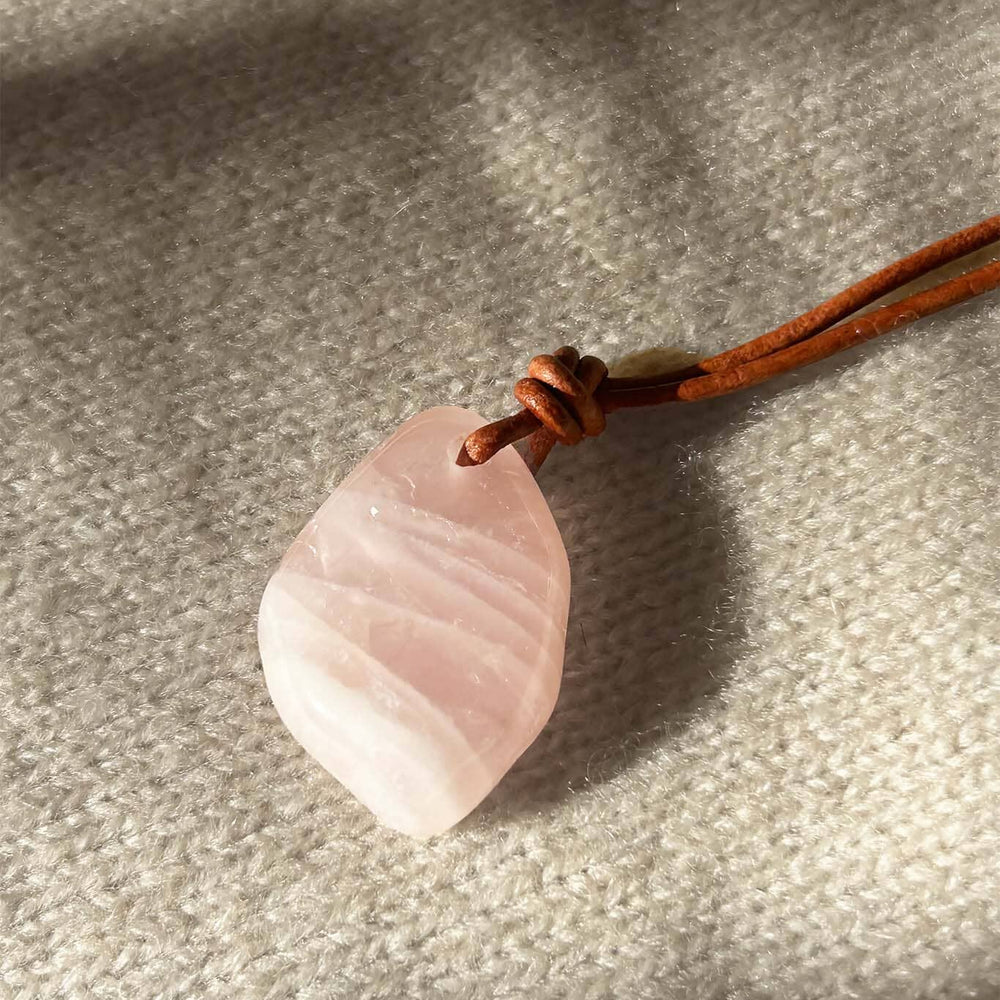 
                  
                    Pink rose quartz stone pendant on a brown cord against a gray fabric background
                  
                