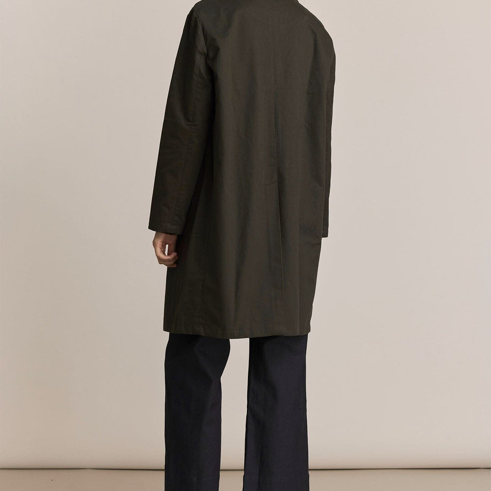 Woman showing the back of a forest green trench coat and black pants against a plain background
