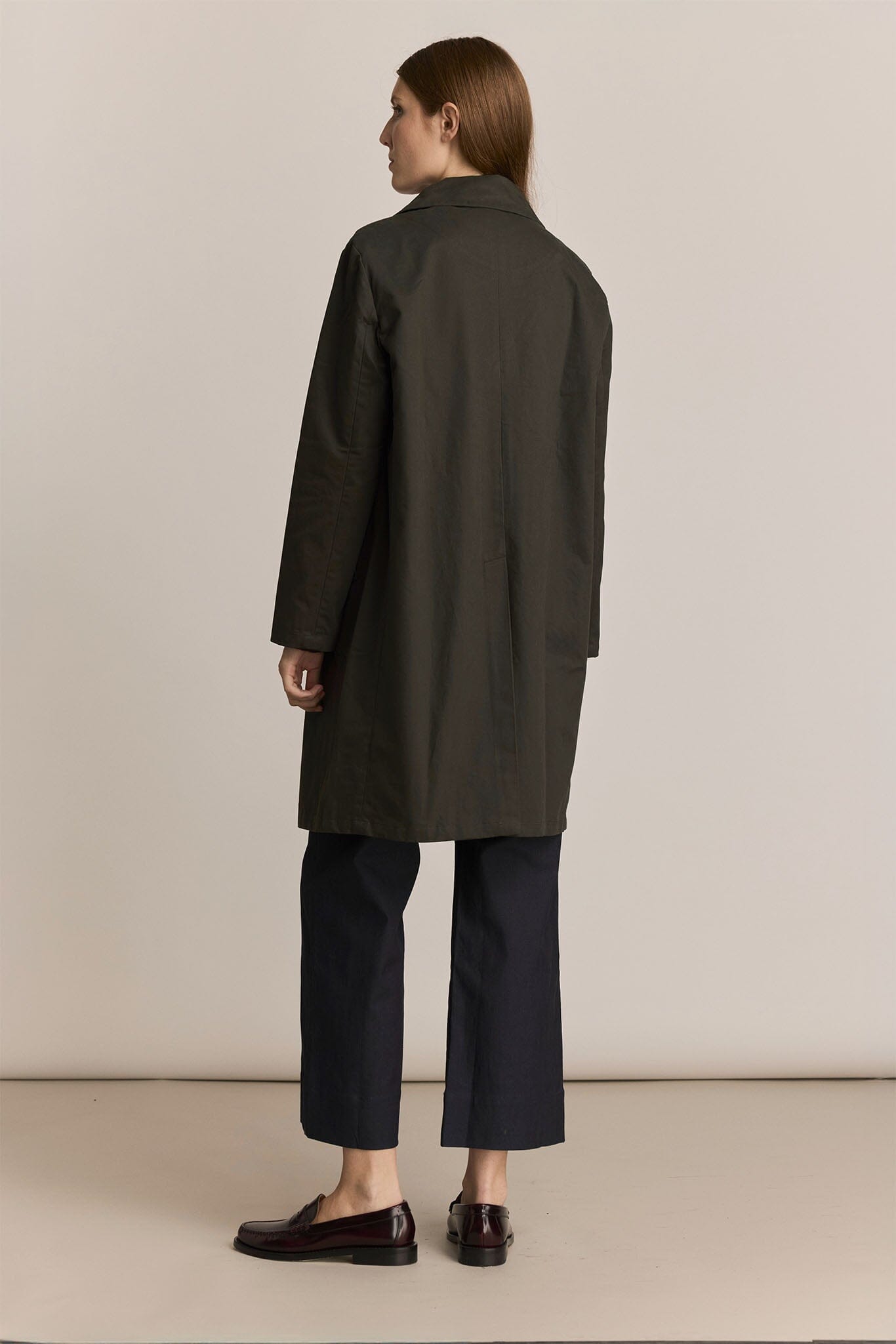 Woman showing the back of a forest green trench coat and black pants against a plain background