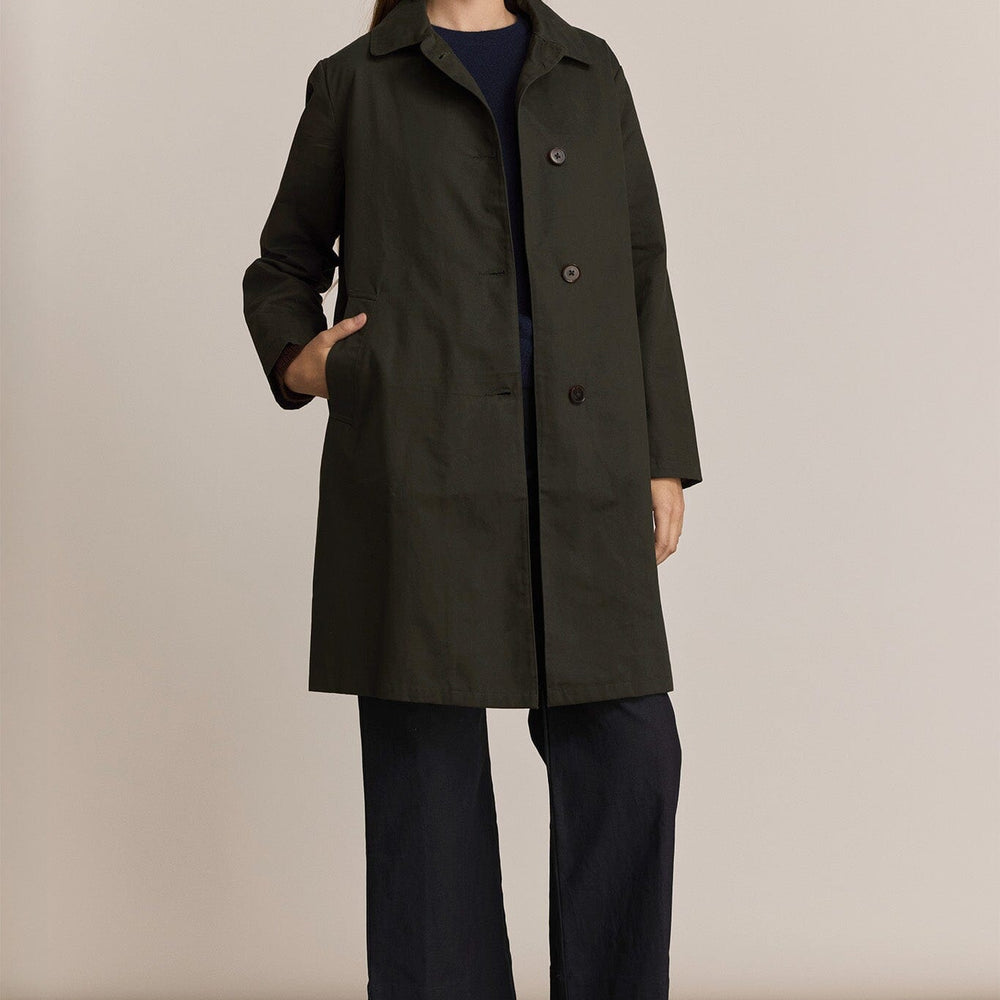 Woman wearing a dark green trench coat and black pants against a plain background