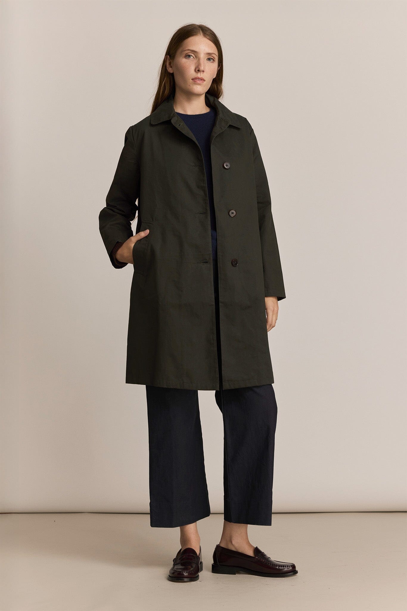 Woman wearing a dark green trench coat and black pants against a plain background