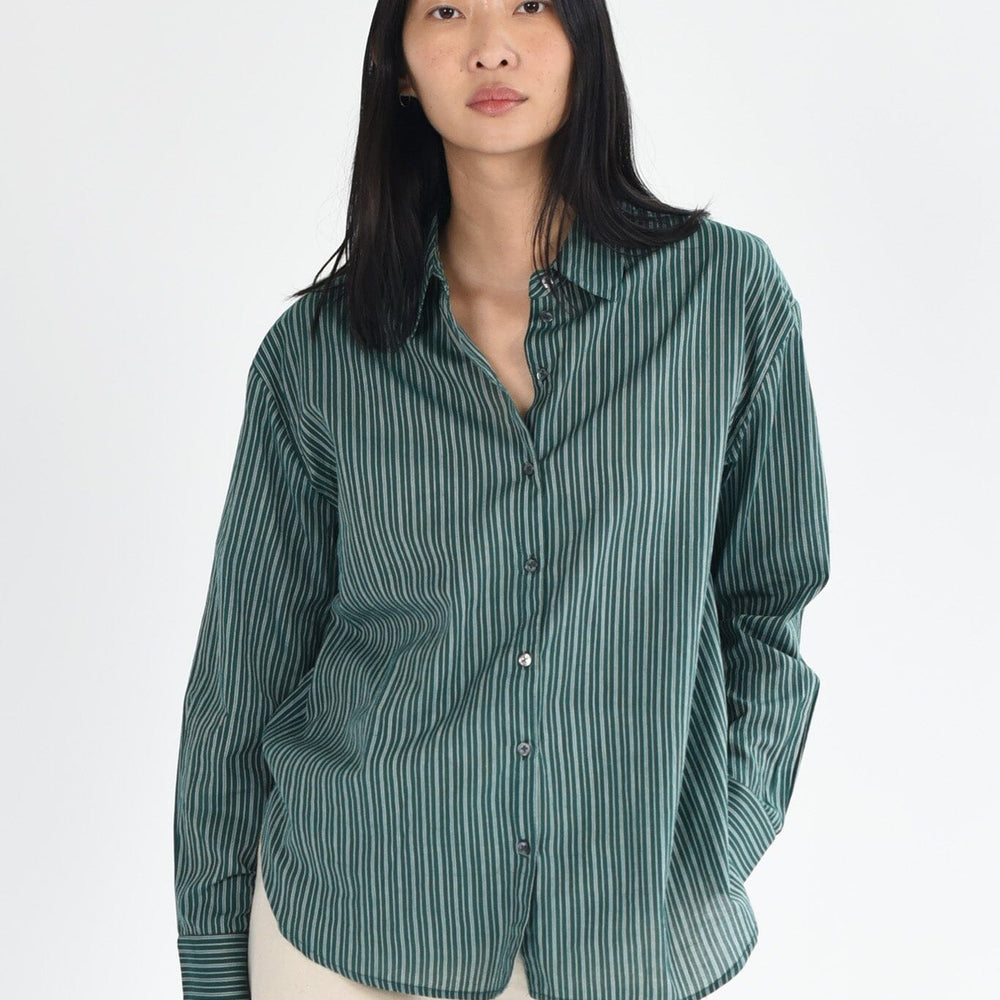 A woman wears a dark green, striped long sleeve button down blouse. The top is lightweight and made of cotton. Available to purchase at M.Patmos in Brooklyn, New York. 