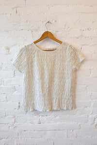 Textured beige short sleeve top on a wooden hanger against a white brick wall