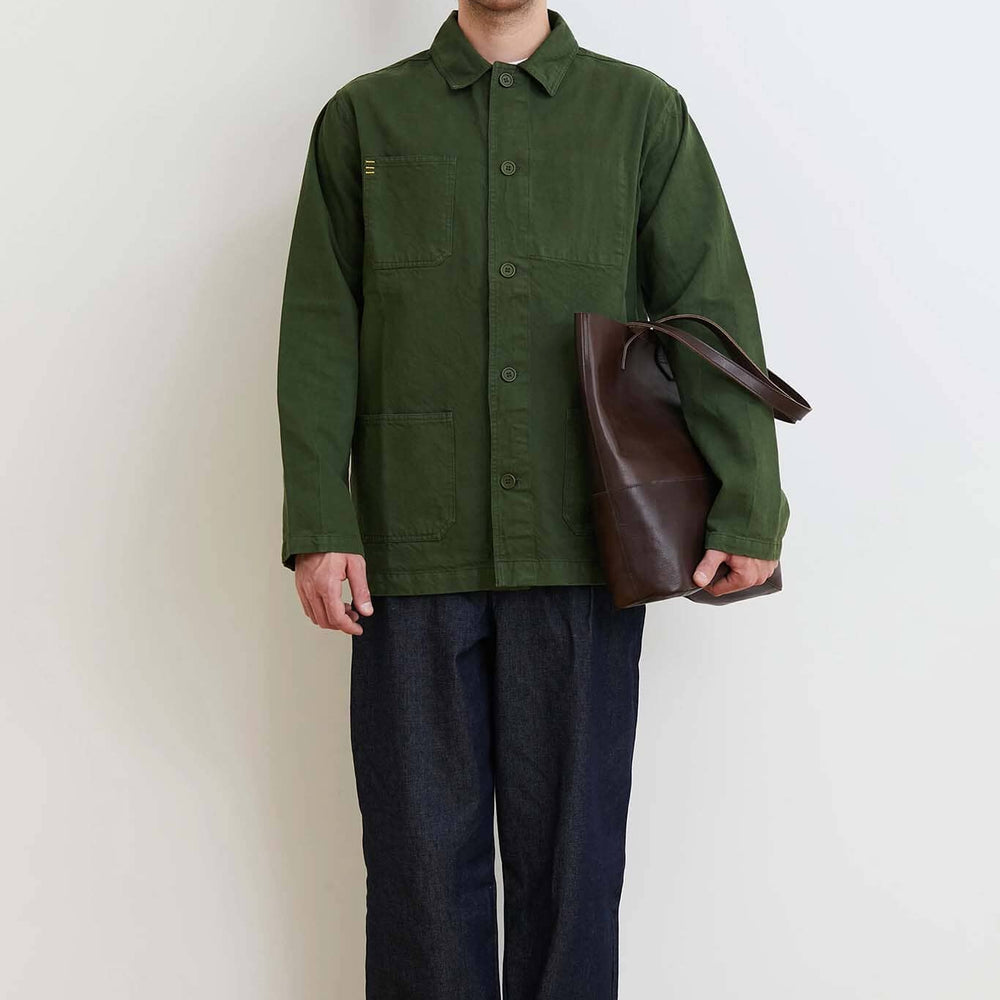 Man wearing a green jacket and dark pants holding a brown bag against a white wall.