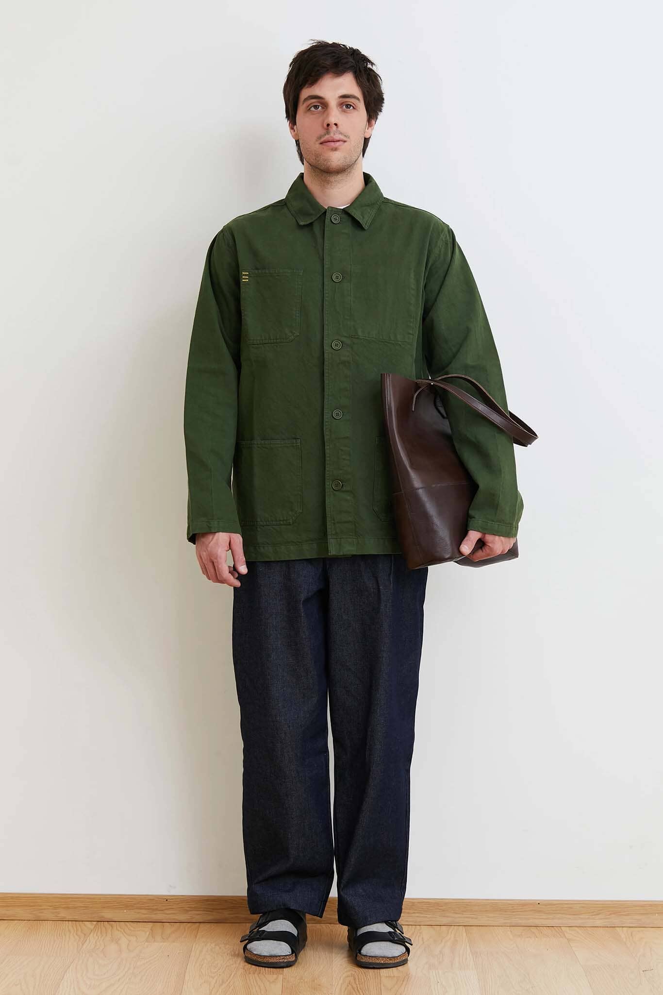 Man wearing a green jacket and dark pants holding a brown bag against a white wall.