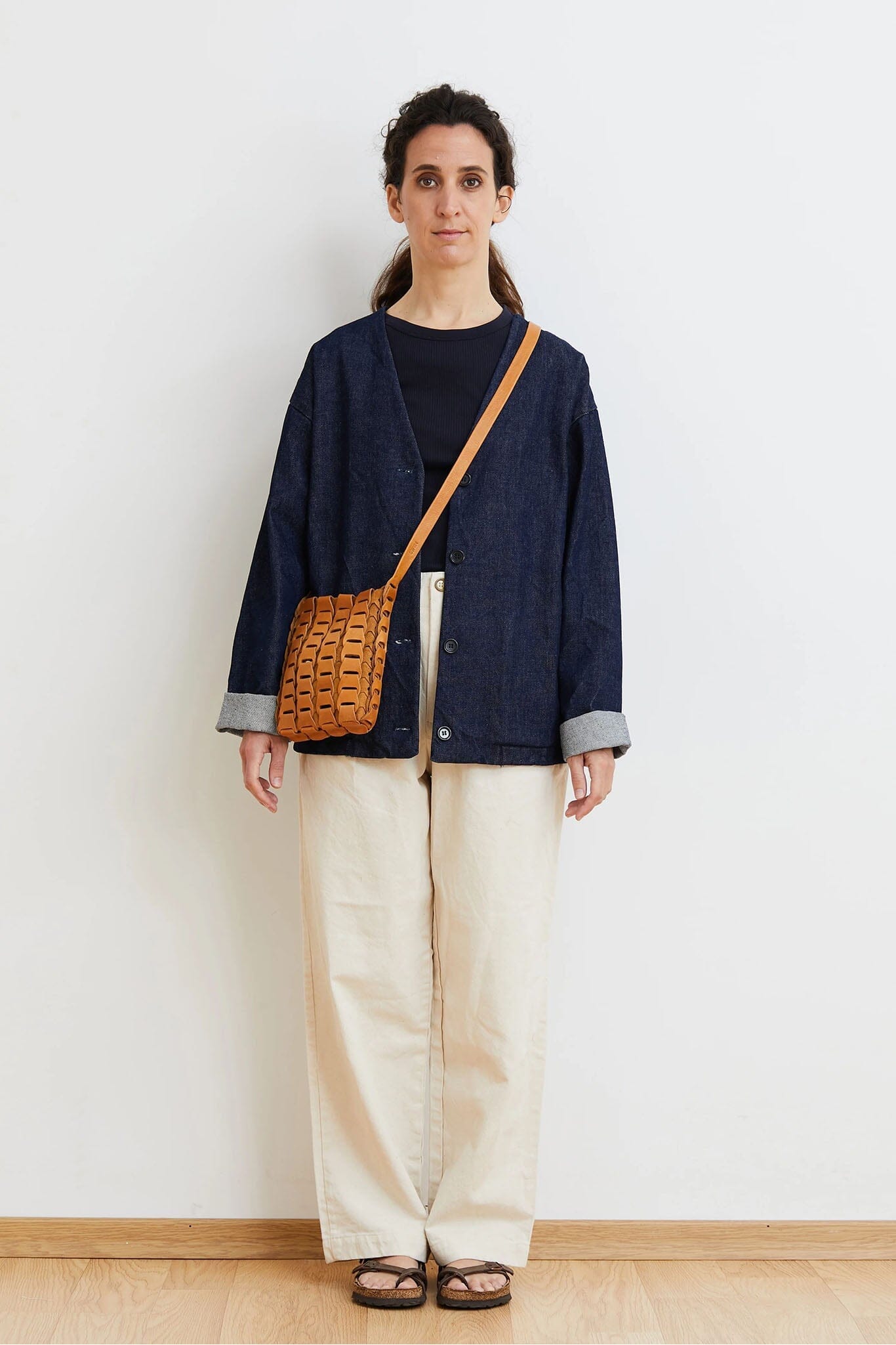 Woman wearing a navy jacket and beige pants with a brown bag against a white wall.