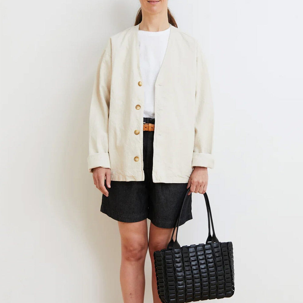Woman wearing a cream cardigan, white shirt, black shorts, and sandals, holding a black woven handbag against a white background.