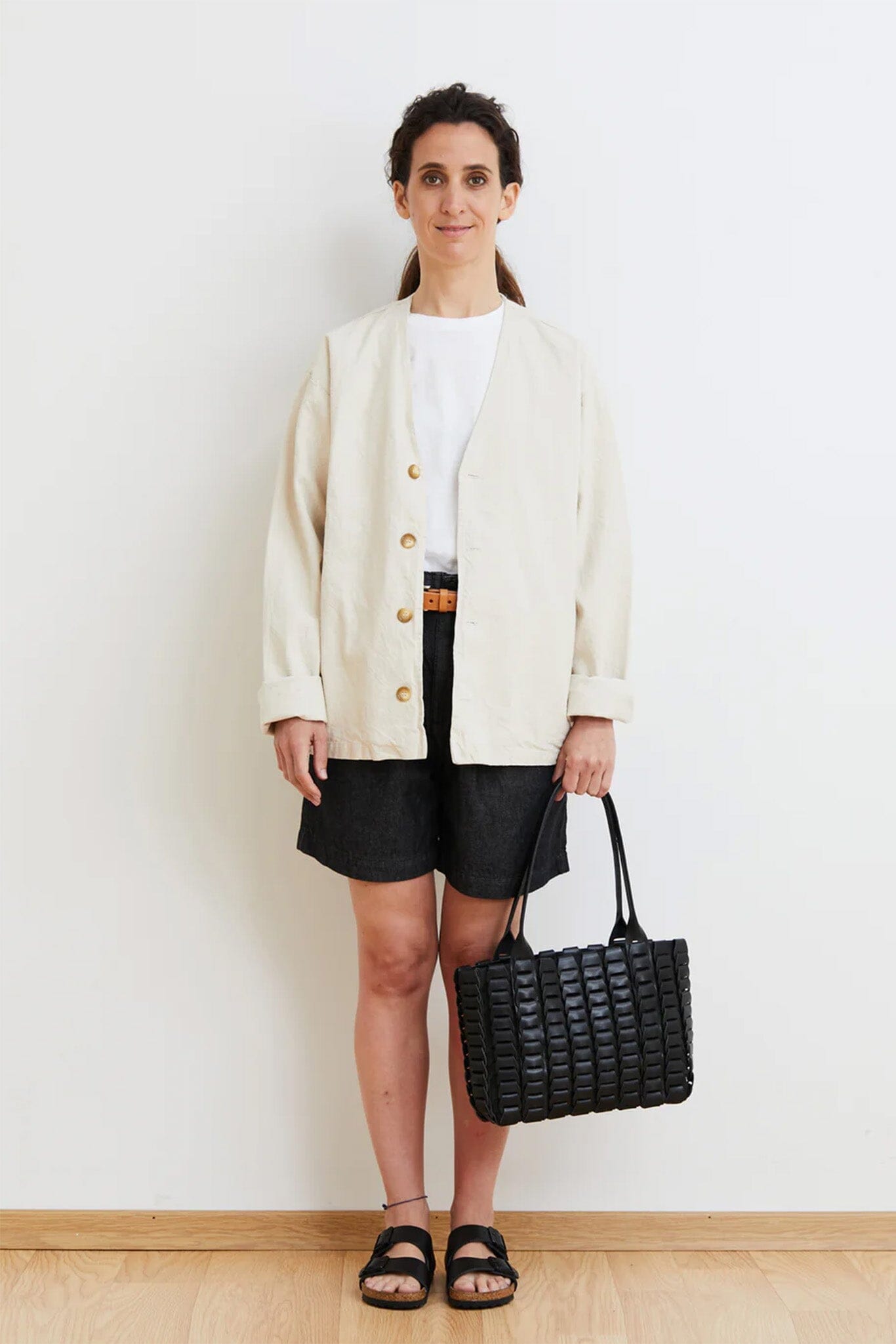 Woman wearing a cream cardigan, white shirt, black shorts, and sandals, holding a black woven handbag against a white background.