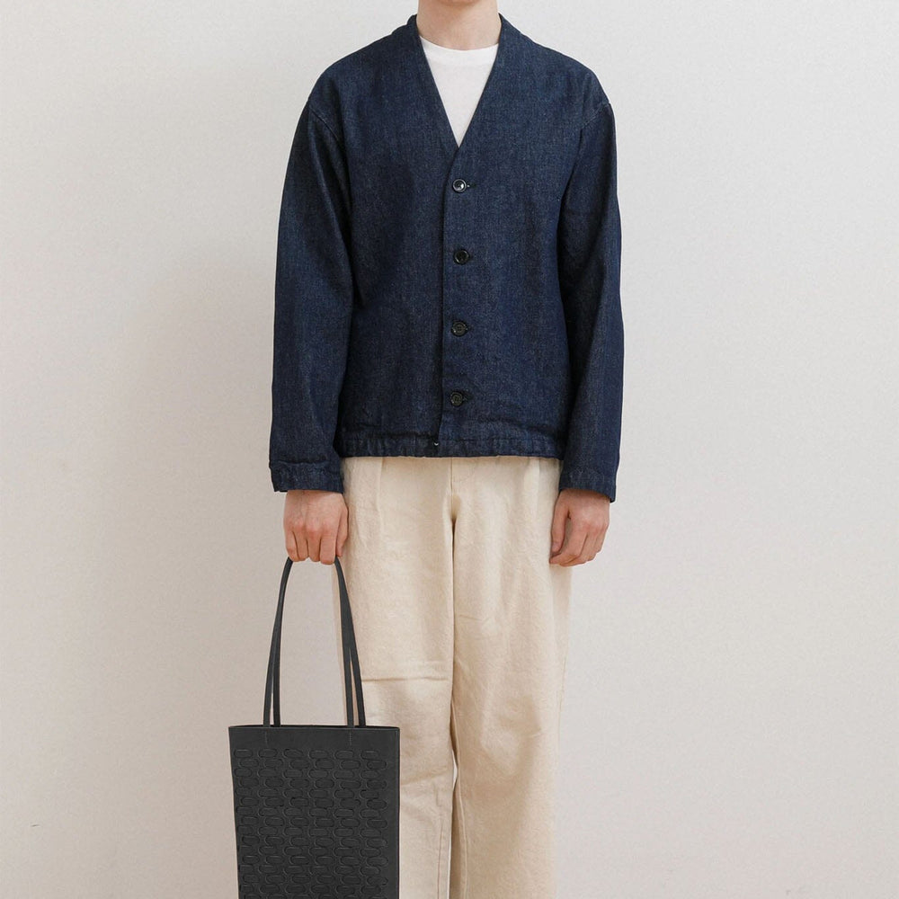 Man wearing a navy cardigan, white shirt, beige pants, and black shoes, holding a black woven tote bag against a plain background.